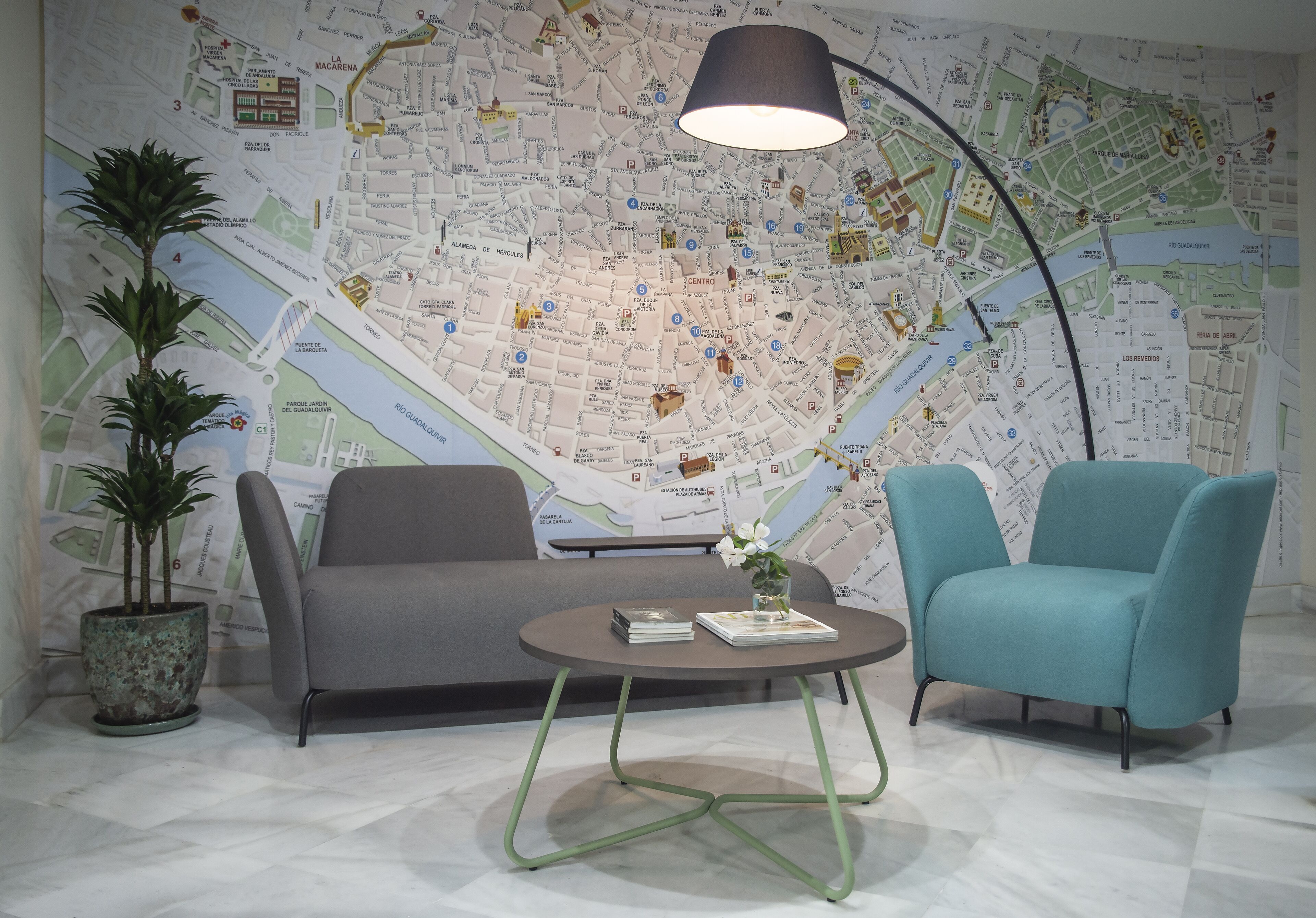 Een kamer met een grote kaart aan de muur, een grijze bank, een blauwgroene fauteuil, een ronde tafel met een lamp en een potplant.