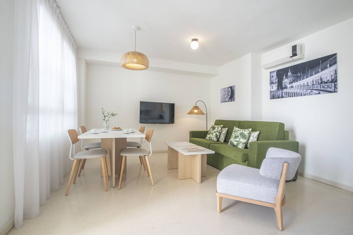 A modern living room with a dining area, a green sofa, and a flat-screen TV mounted on the wall.