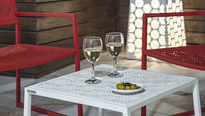 Una mesa blanca con una copa de vino blanco y un bol de aceitunas, sillas rojas y un panel de luz decorativo en un jardín.