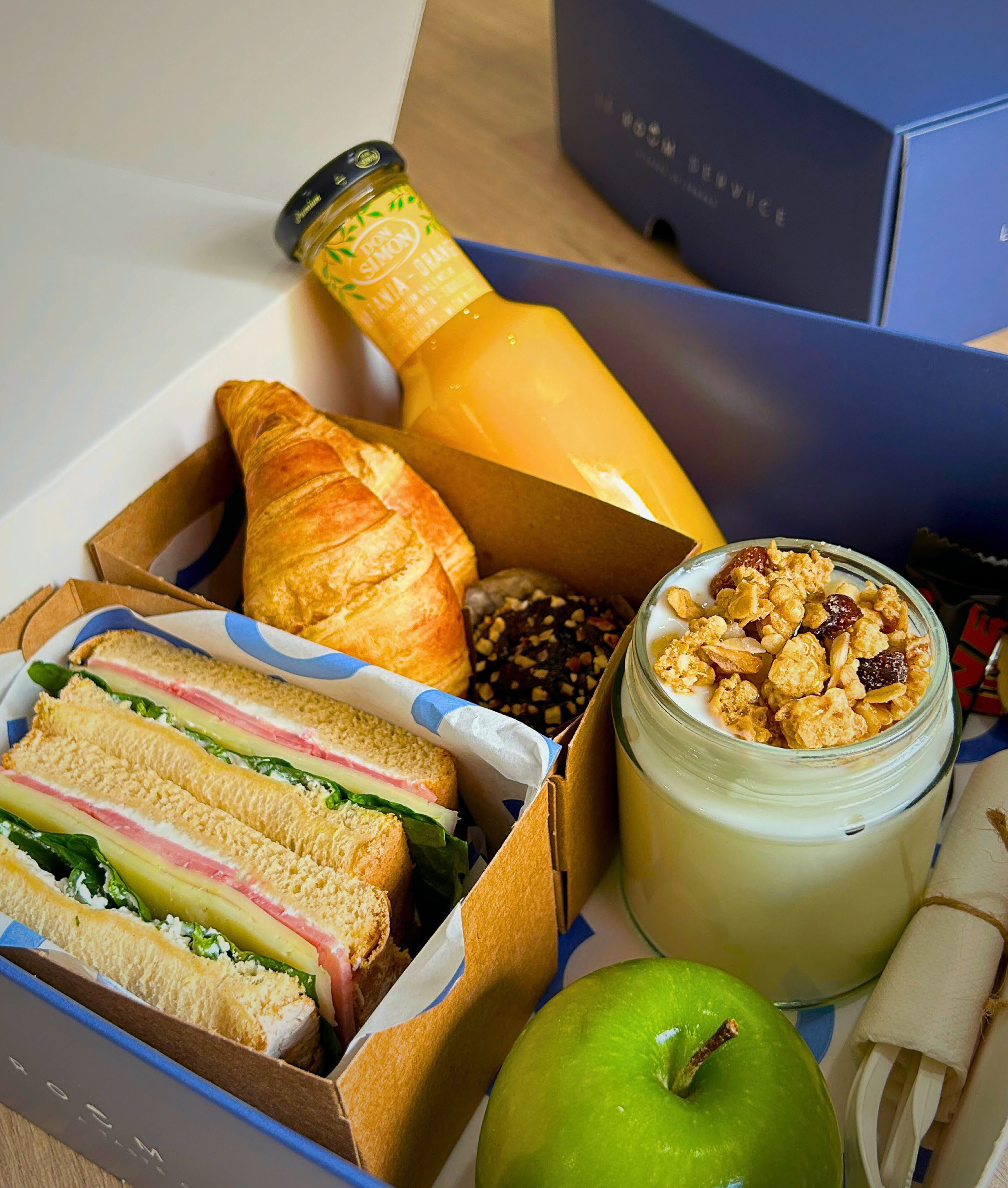 Una caja con un sándwich, un croissant, una botella de jugo, un tarro de yogur con granola y una manzana.