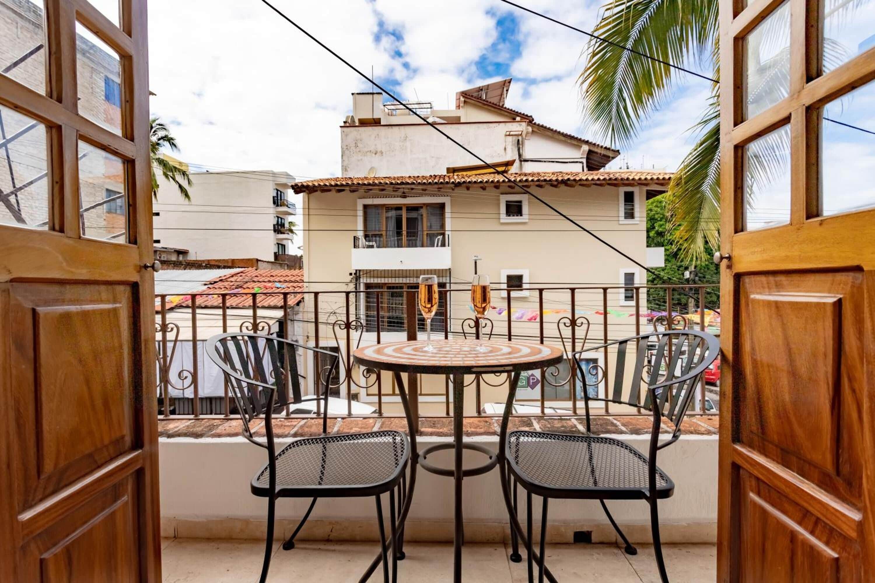 A balcony with two metal chairs and a small round table, overlooking a residential area with a tiled roof building.