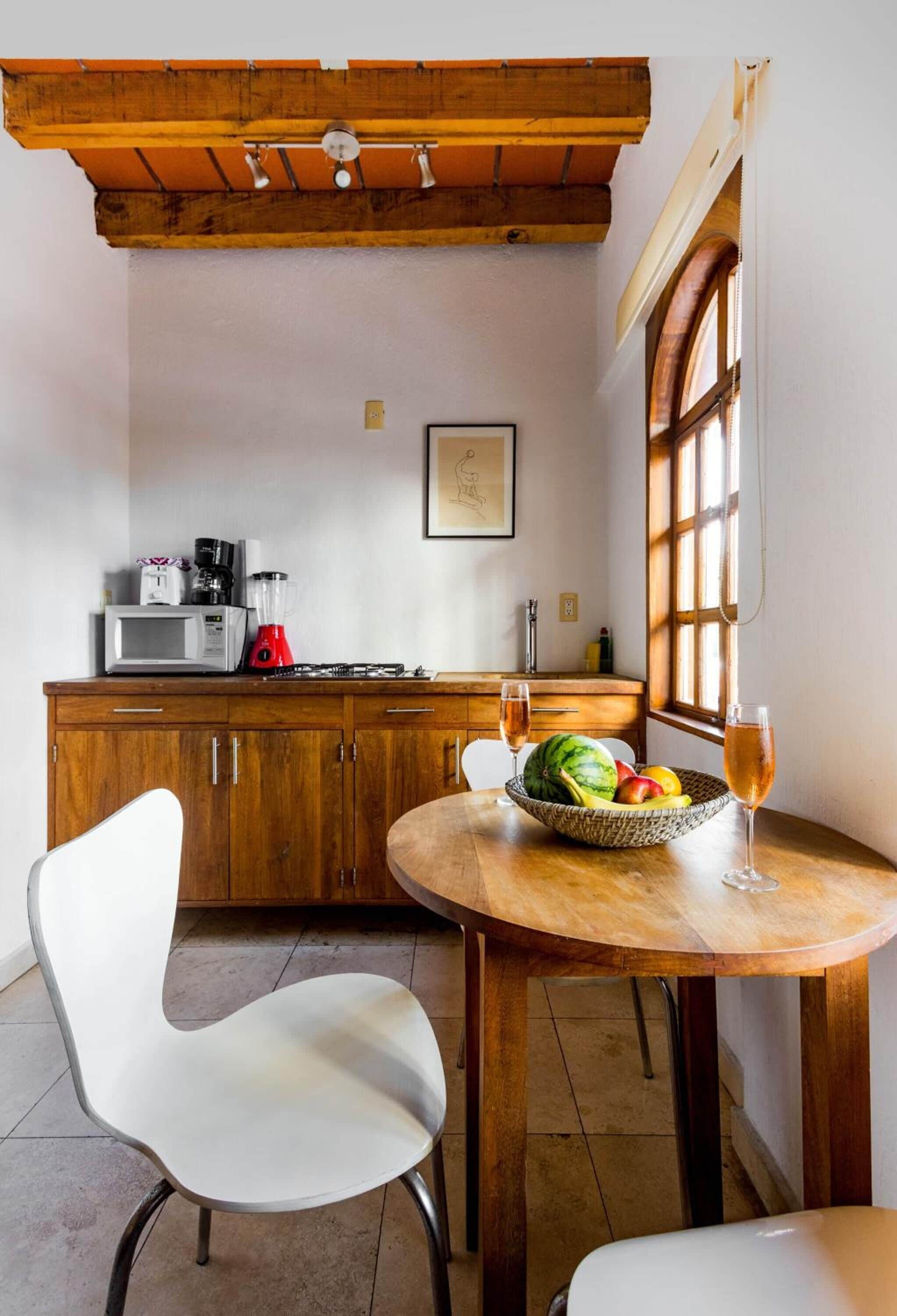 A kitchen with wooden cabinets, a microwave, a blender, and a fruit bowl on a table with a glass of wine.