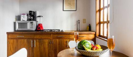 A kitchen with wooden cabinets, a microwave, a blender, and a fruit bowl on a table with a glass of wine.