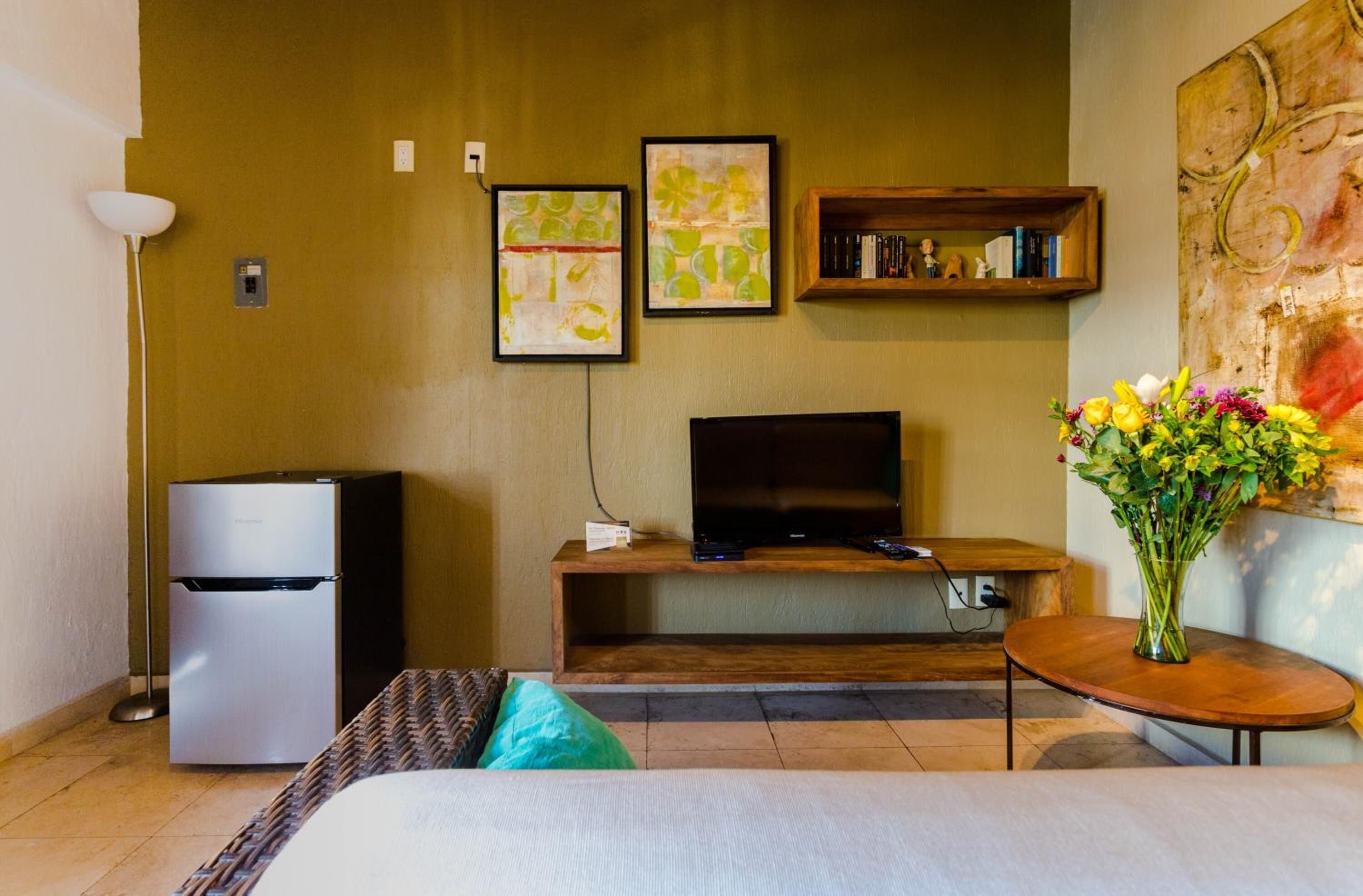 A hotel room with a bed, a television on a wooden stand, a refrigerator, a small table with a vase of flowers, and artwork on the wall.