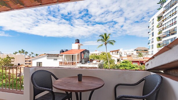 A balcony with two wicker chairs and a small round table, overlooking a cityscape with buildings and a palm tree. - Olas Altas Suites (Puerto Vallarta)