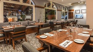 A restaurant with a long wooden table set for dining, multiple seating areas including a sofa and chairs, and a bar area visible through large windows. - Olas Altas Suites (Puerto Vallarta)