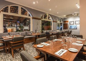 Un restaurante con una larga mesa de madera lista para comer, múltiples zonas de estar que incluyen un sofá y sillas, y un área de bar visible a través de grandes ventanales.