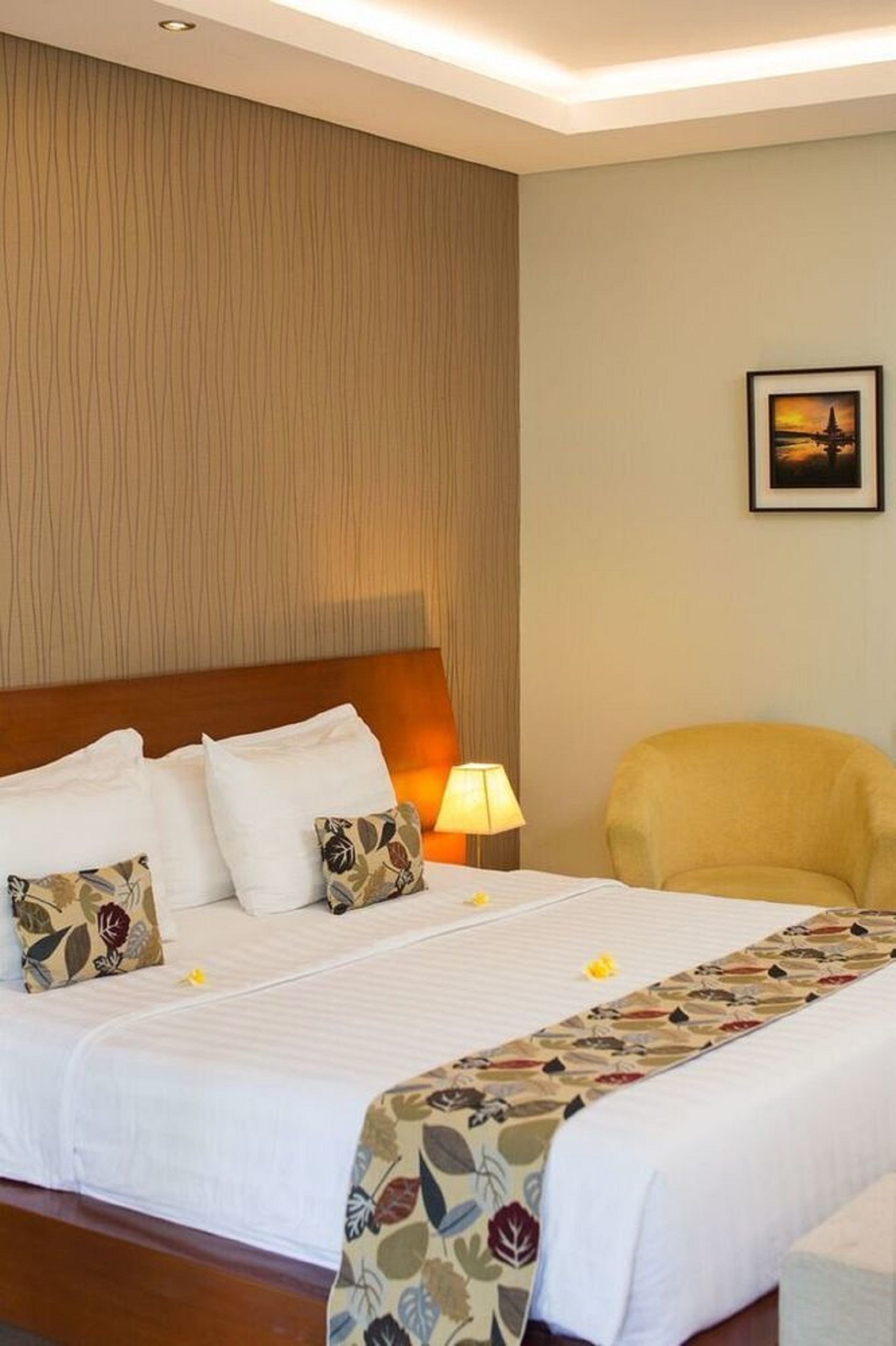 A hotel room with a large bed, a wooden headboard, a bedside lamp, a chair, and a framed picture on the wall.