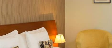 Habitación de hotel con una cama grande, cabecera de madera, lámpara de noche, una silla e imagen enmarcada en la pared.