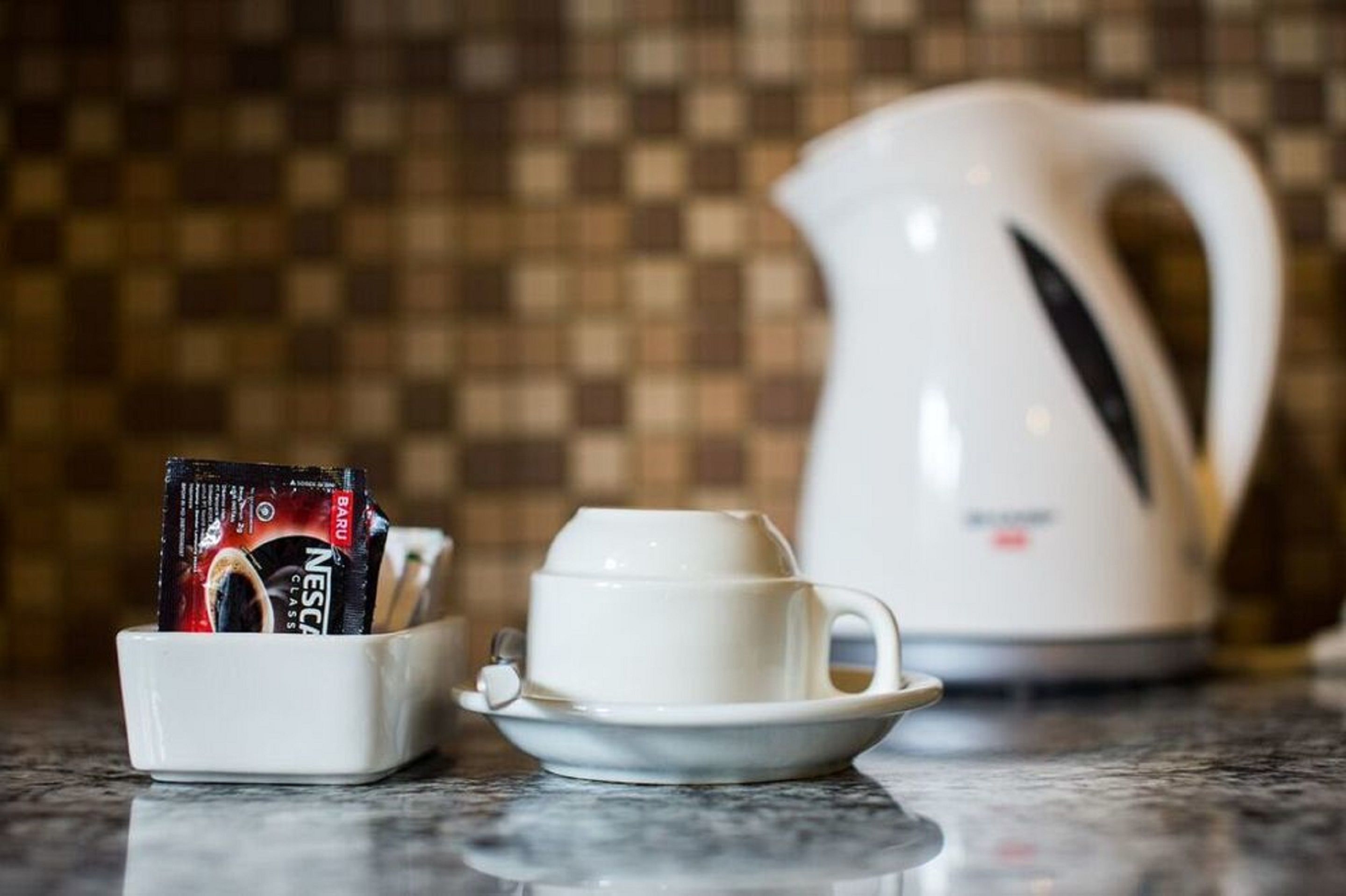 A white coffee cup and saucer on a marble countertop, a white electric kettle, and a coffee pod holder with a Nescafé pod.