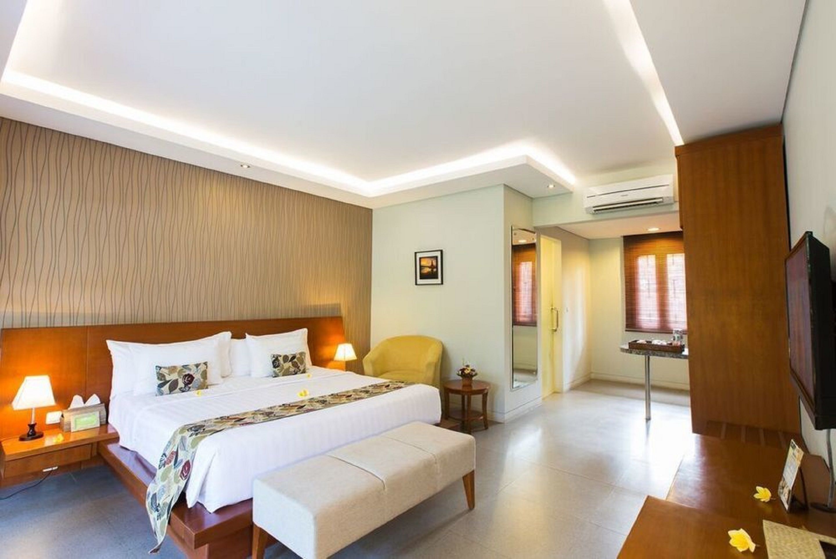 Quarto de hotel com uma cama grande, abajures, cabeceira de madeira, uma pequena mesa com luminária, uma cadeira e uma televisão.