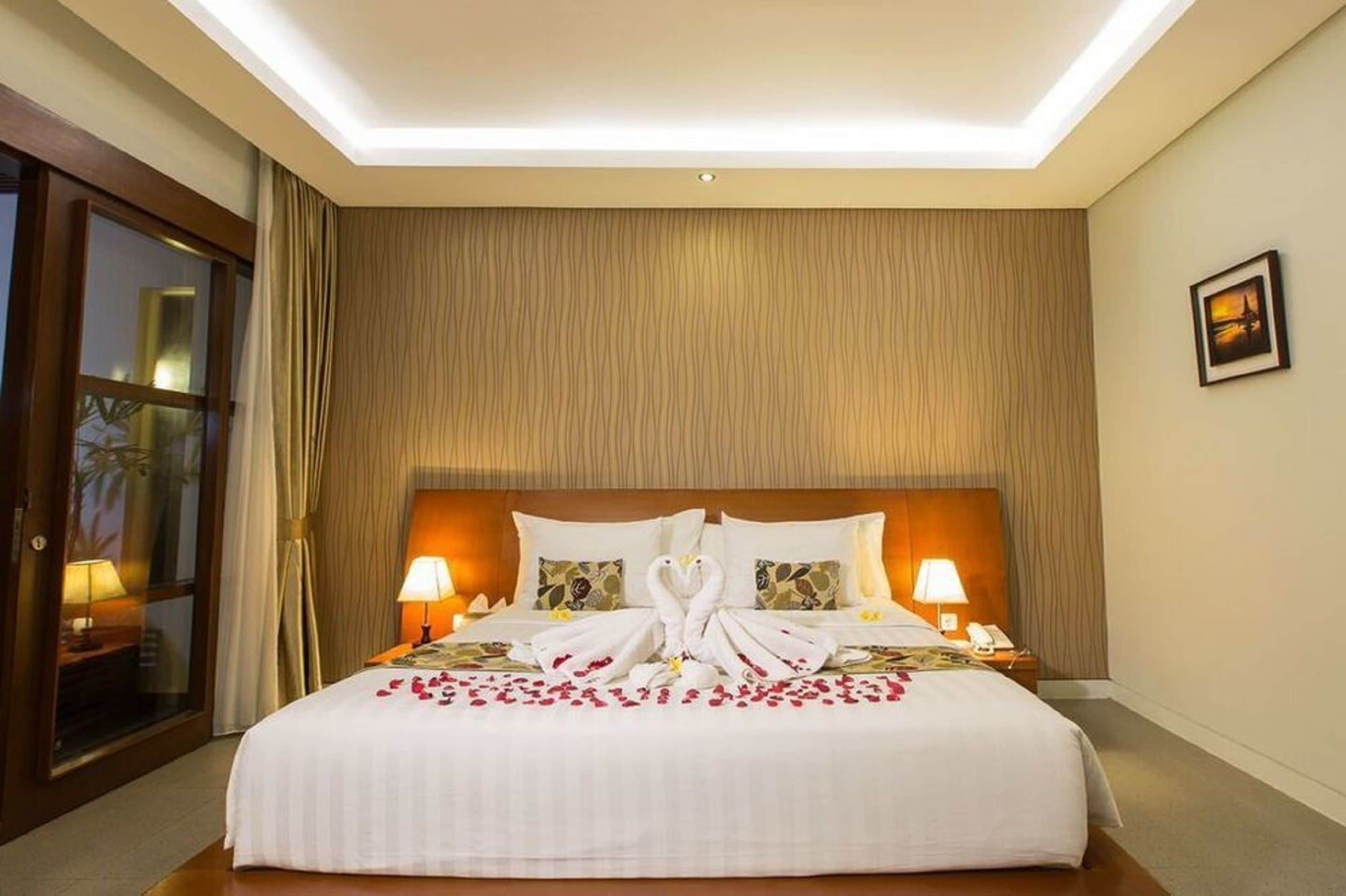 A hotel room with a large bed, two bedside lamps, a wooden headboard, a window with curtains, and a framed picture on the wall.
