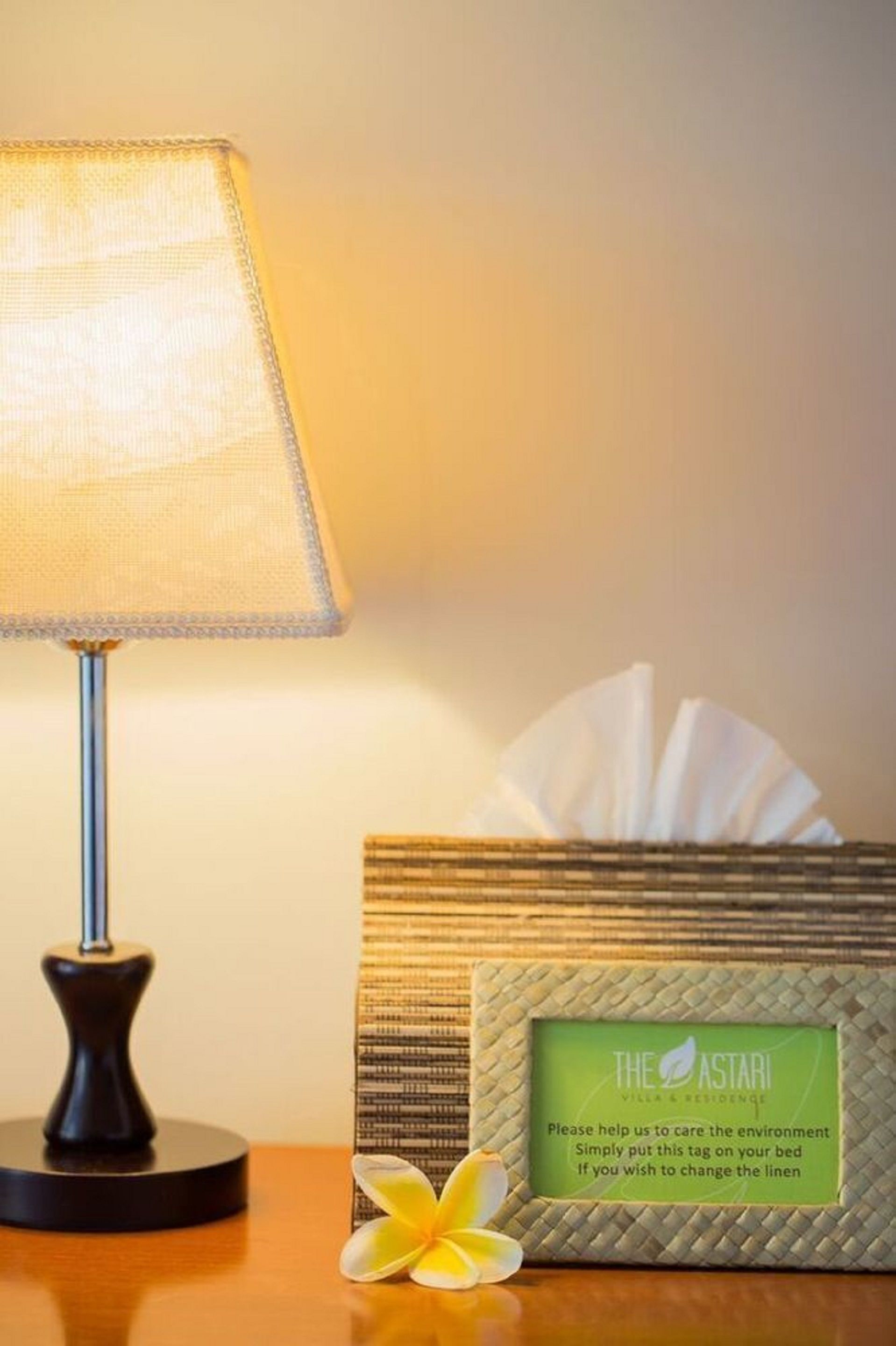A bedside table with a lamp, a tissue box, and a tag instructing to help care for the environment.