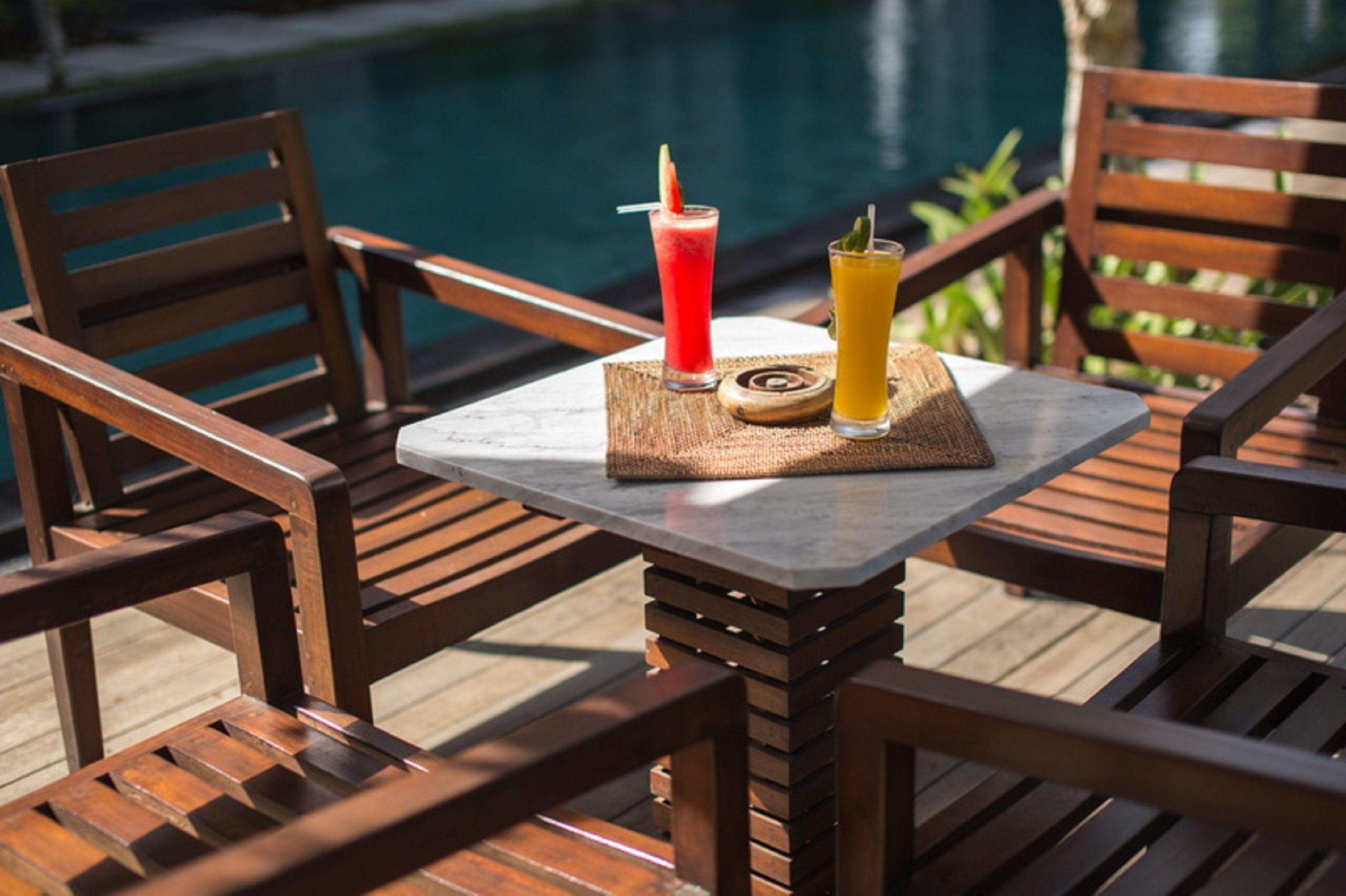 Een houten terras met een tafel en stoelen, twee drankjes op een onderzetter, en een zwembad op de achtergrond.