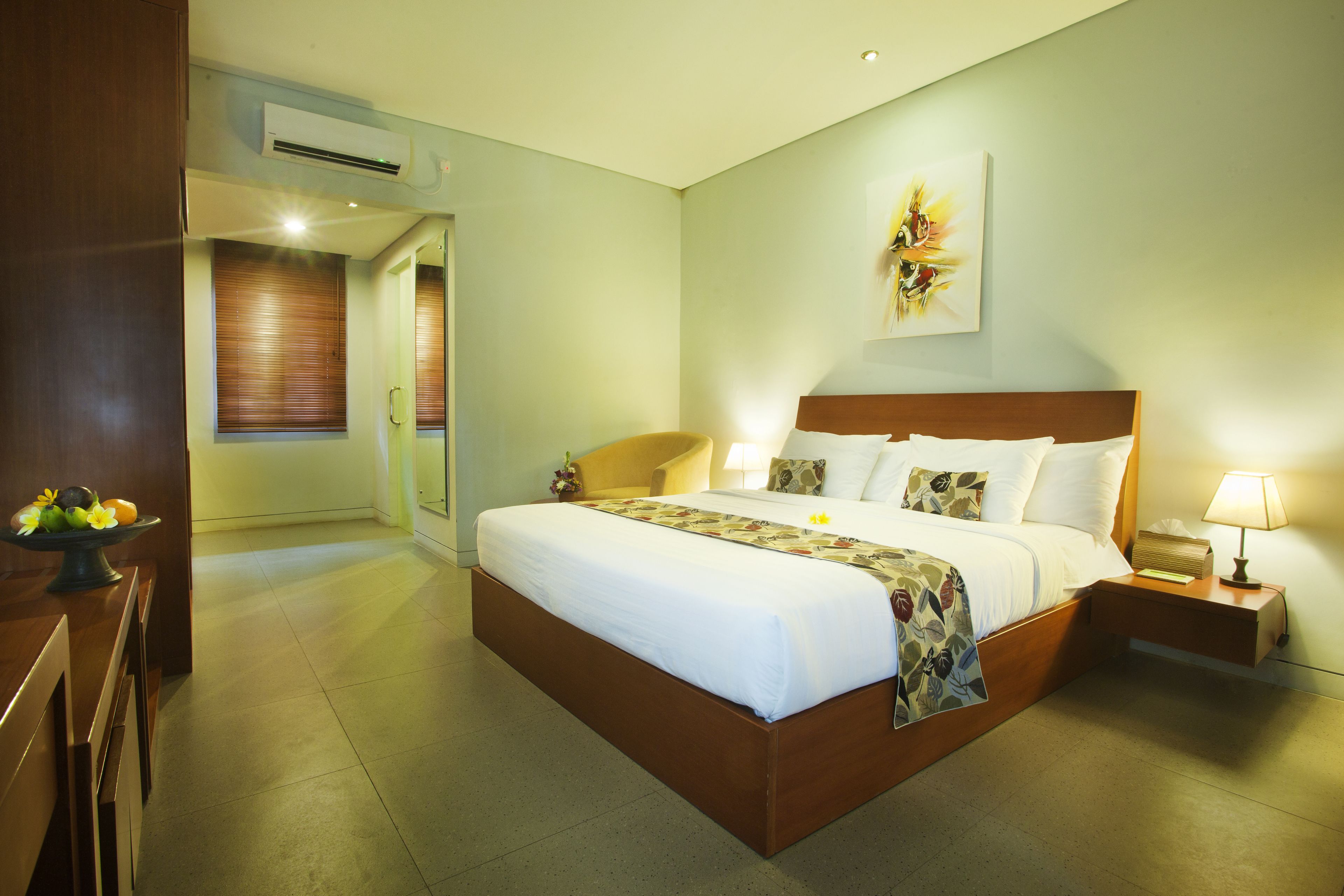 A hotel room with a large bed, bedside tables, an air conditioning unit, a wall-mounted artwork, and a wooden bench.