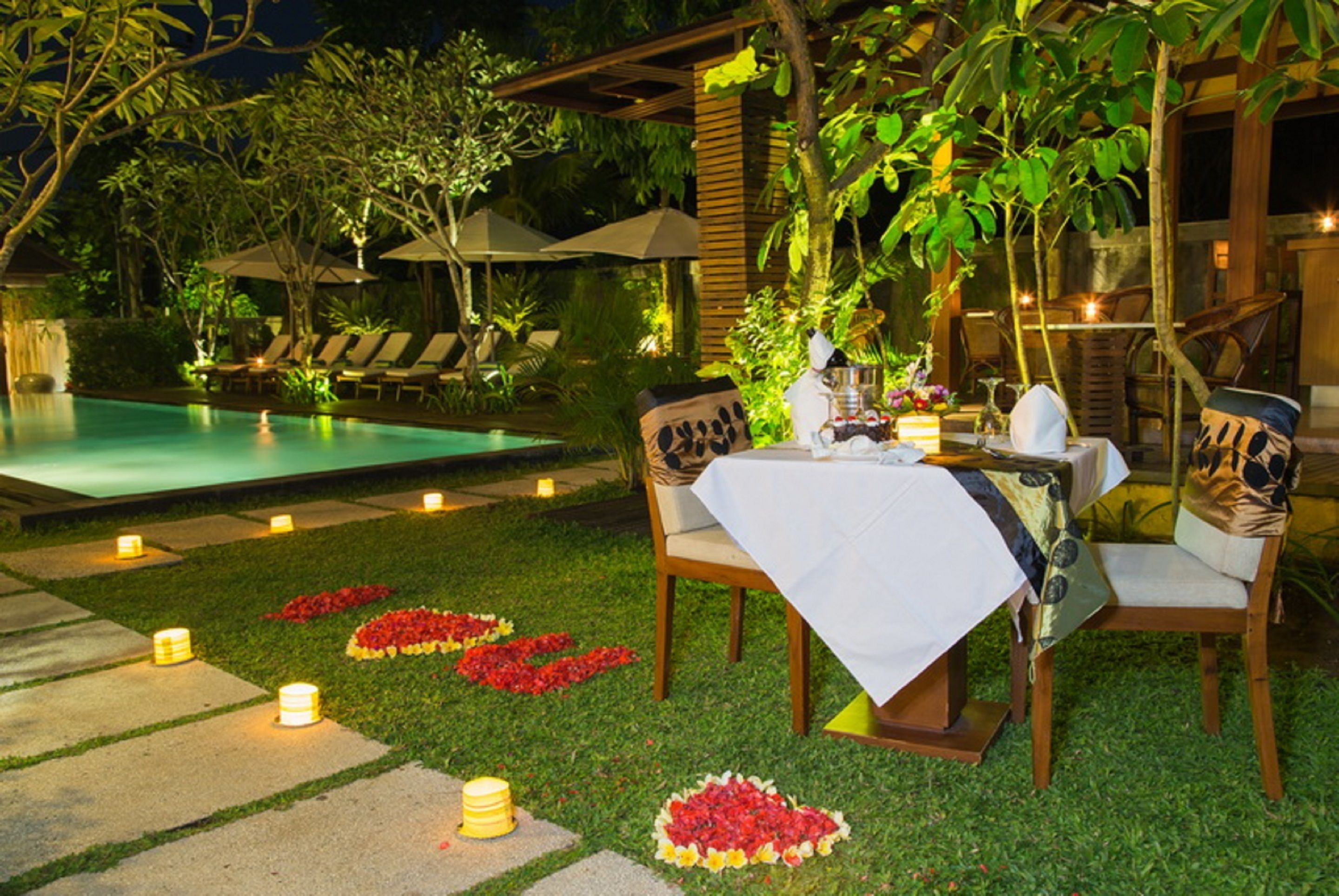 An outdoor dining area with a table set for two, surrounded by chairs, near a pool and garden.