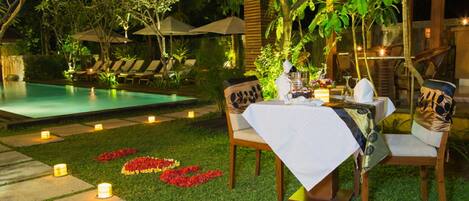 An outdoor dining area with a table set for two, surrounded by chairs, near a pool and garden.
