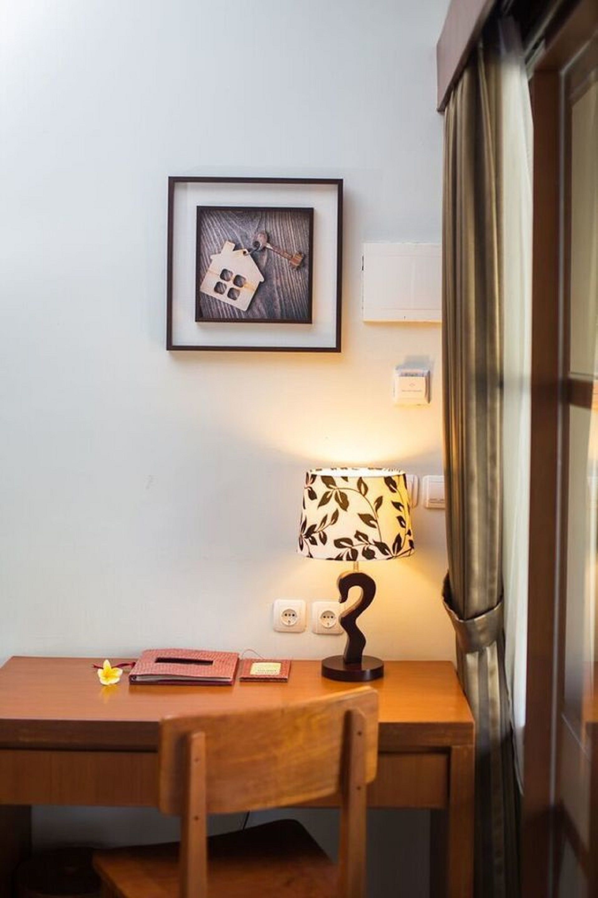 Una habitación pequeña y ordenada con un escritorio de madera, un cuadro en la pared, una lámpara con motivos de hojas y una ventana con cortinas.
