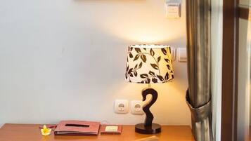 Una habitación pequeña y ordenada con un escritorio de madera, un cuadro en la pared, una lámpara con motivos de hojas y una ventana con cortinas.
