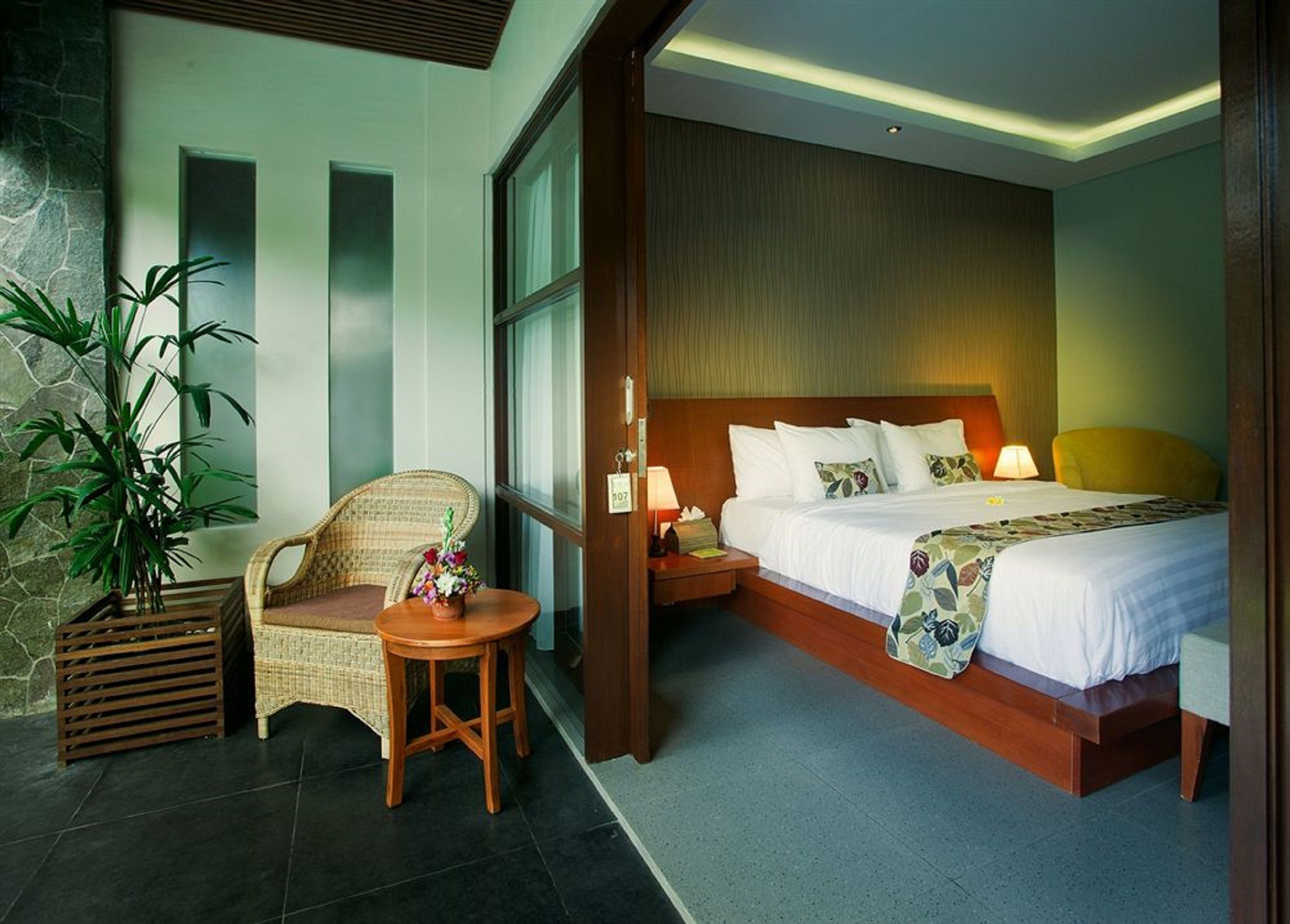 Une chambre d’hôtel avec un lit, une table de chevet, une lampe, une chaise et une grande plante en pot.