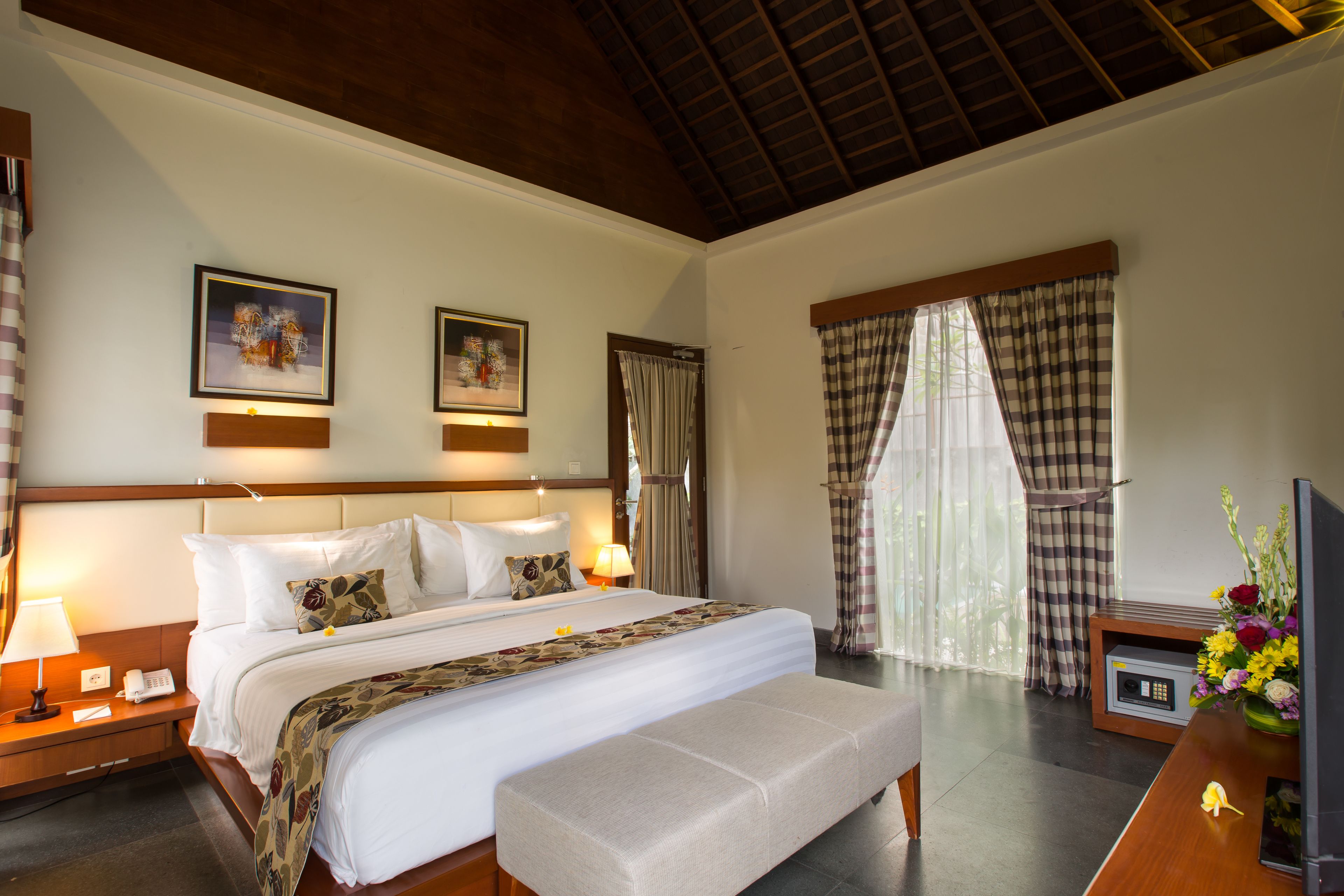 A hotel room with a large bed, bedside tables with lamps, a desk with a TV and flowers, and a window with curtains.