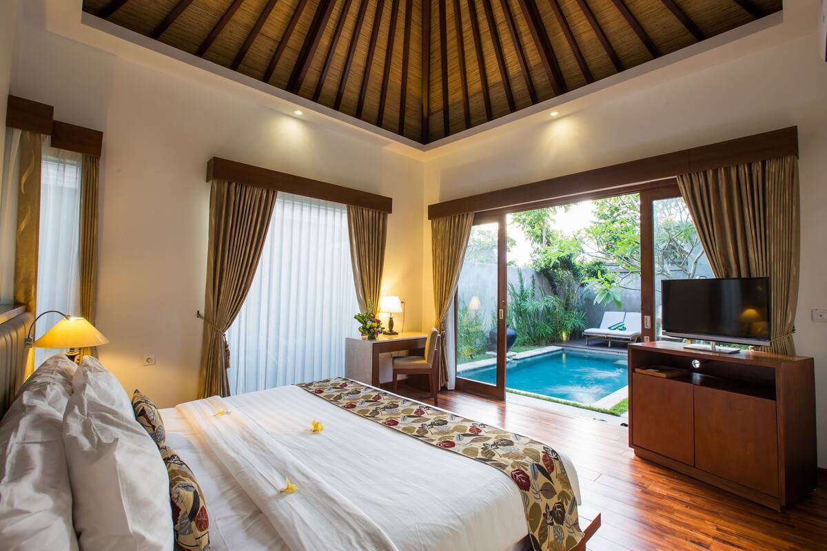 Una camera d'albergo con un letto, una scrivania con una sedia, una televisione e vista su una piscina.