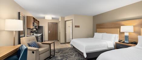 A hotel room with two beds, a desk, a chair, a microwave, a refrigerator, a small table, and a nightstand with a lamp.