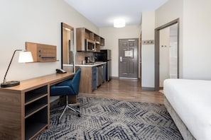 Une chambre d’hôtel compacte comprenant un coin cuisine, un bureau avec une chaise, un lit et un tapis à motifs.