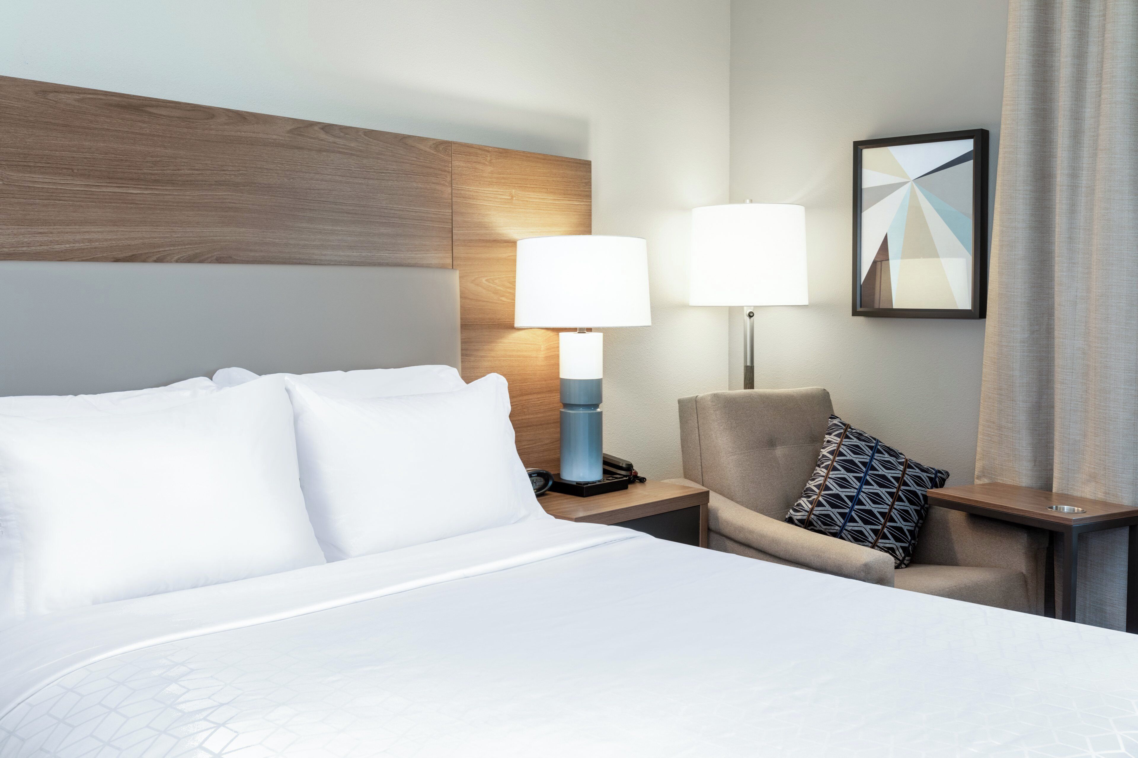 Une chambre d’hôtel avec un lit, une chaise, une table de chevet, une lampe et un tableau encadré au mur.