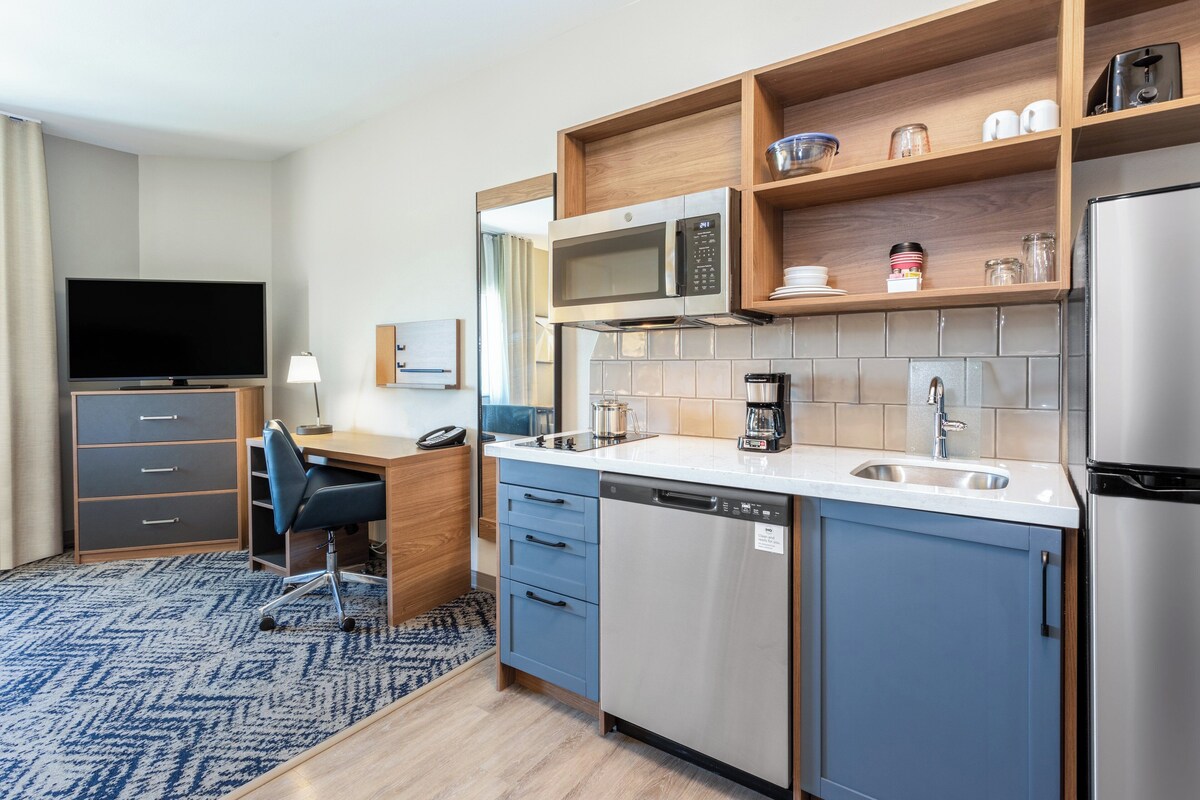 A modern kitchen with a microwave, coffee maker, and dishwasher. There is a TV and a desk with a chair.