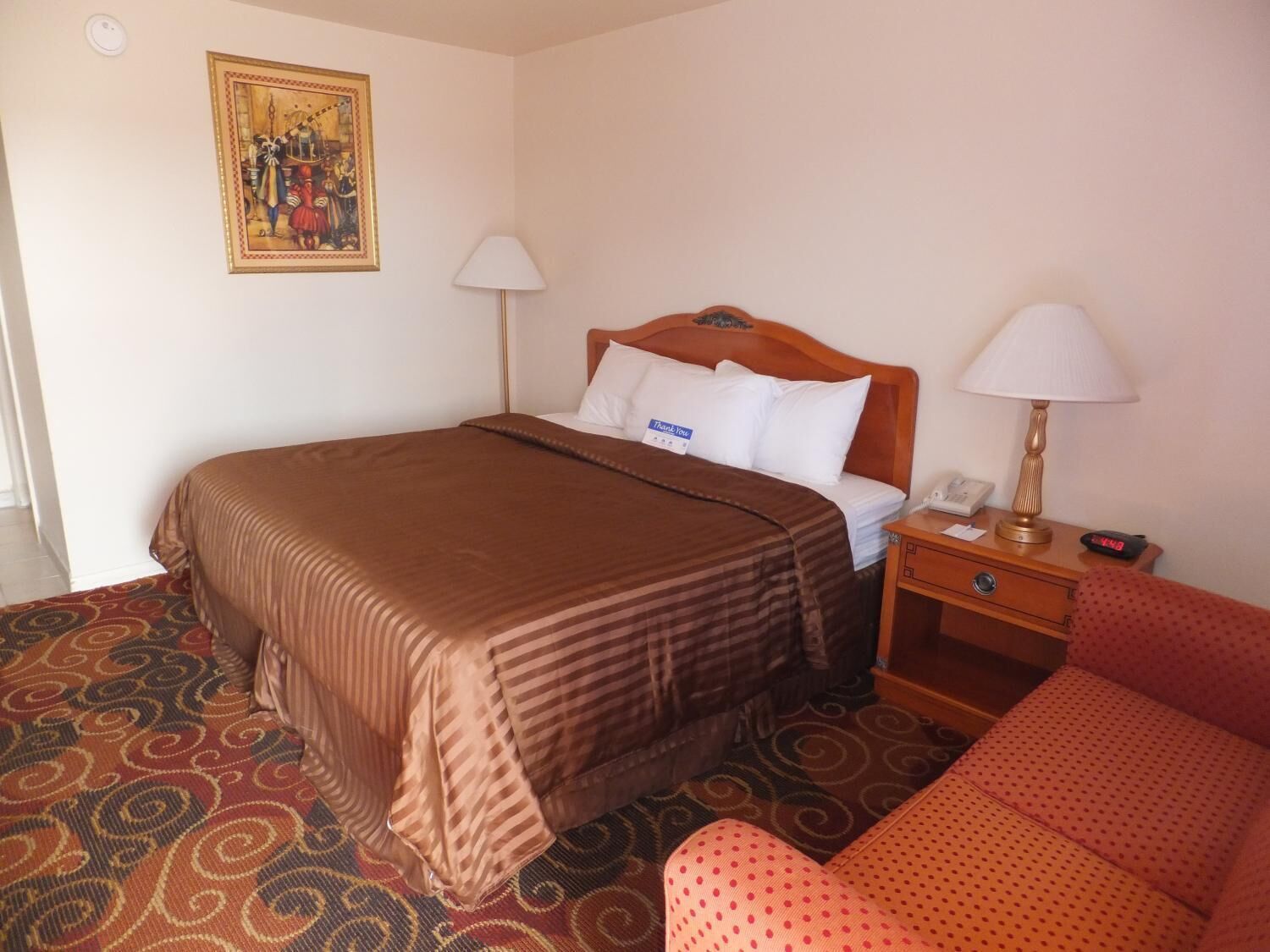 A hotel room with a bed, a nightstand, a lamp, a chair, and a painting on the wall.