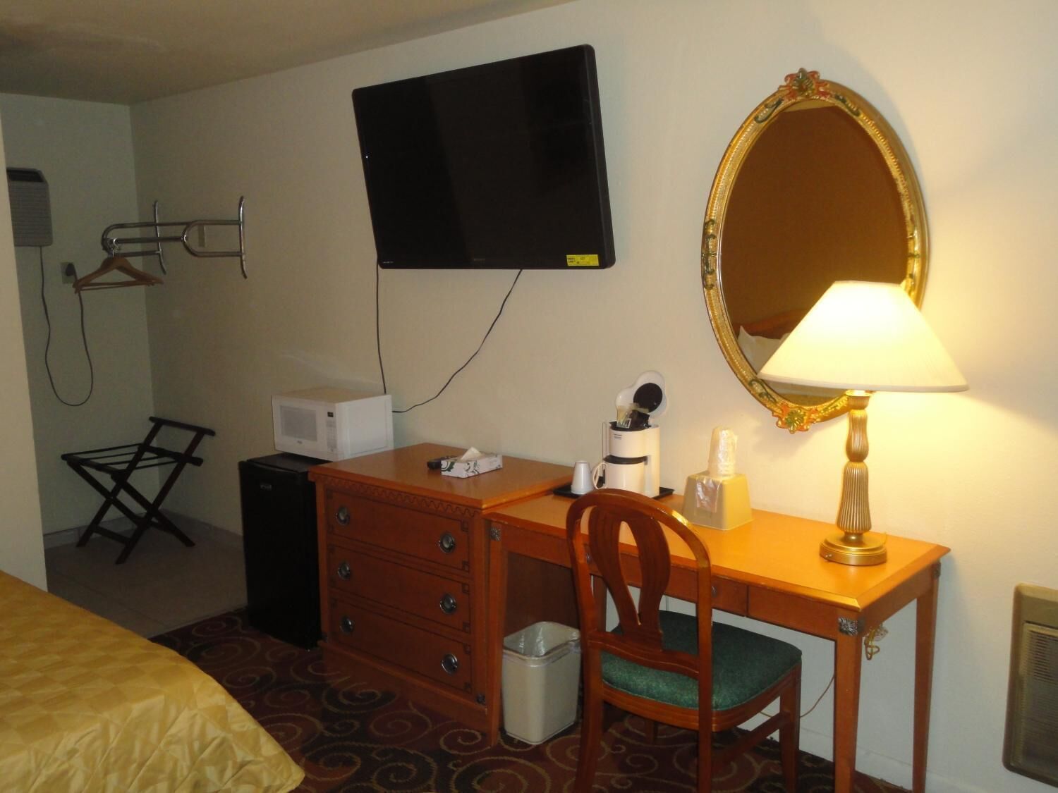 A hotel room with a wooden desk, a television mounted on the wall, a mirror, a lamp, a chair, a microwave, a small trash bin, and a bed with a patterned bedspread.