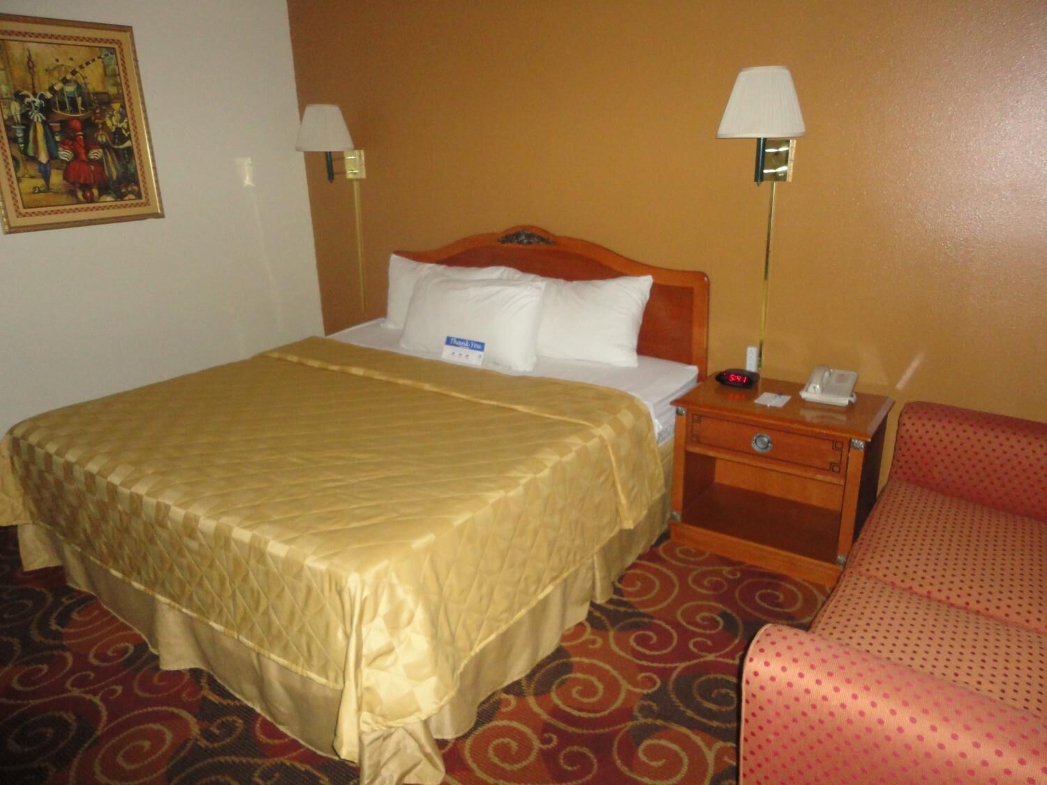 A hotel room with a large bed, two bedside lamps, a nightstand with a phone and a clock, and a patterned carpet.