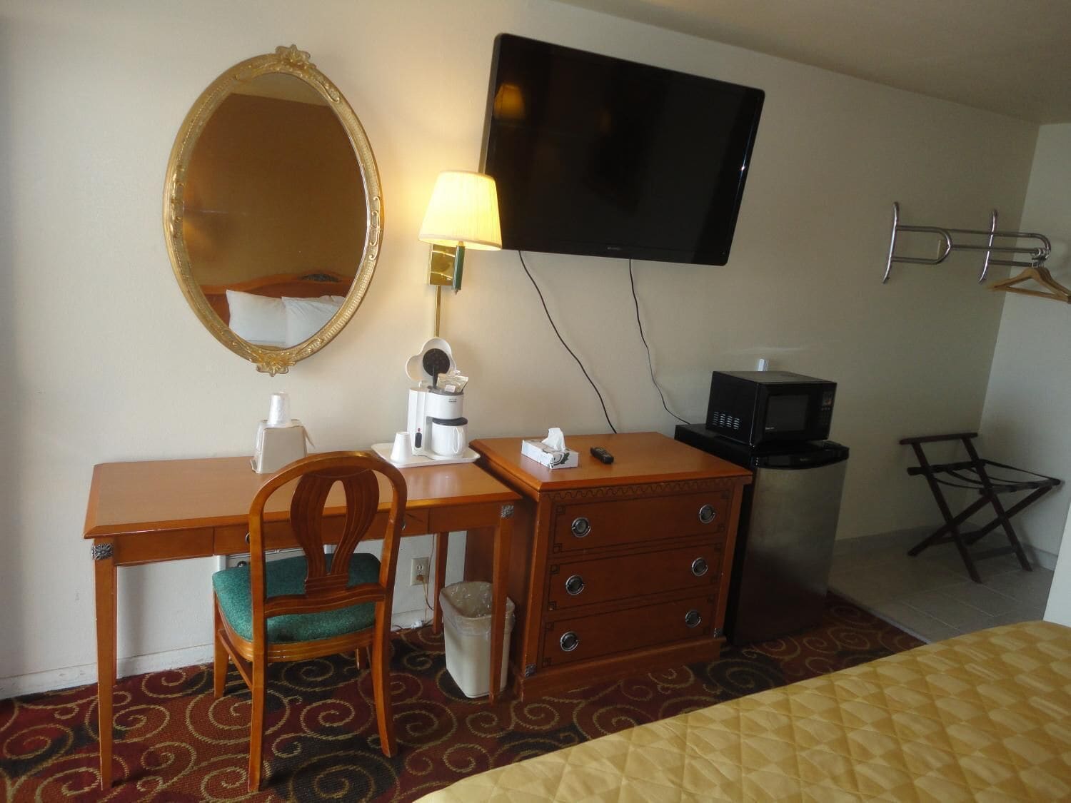 Habitación de hotel con un escritorio de madera, una silla, un televisor montado en la pared, una mesita con una lámpara, un cesto de basura y un perchero.