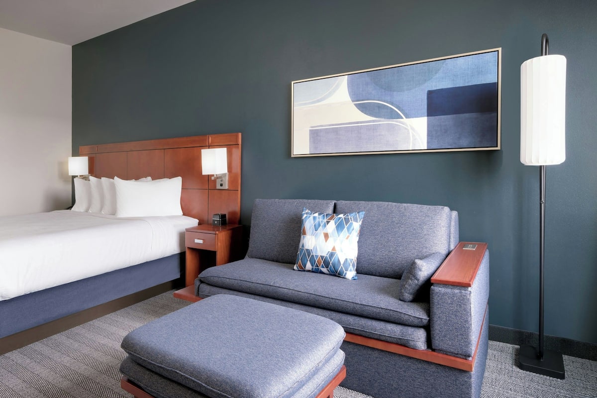 A hotel room with a bed, a sofa, a nightstand, a lamp, and a painting on the wall.