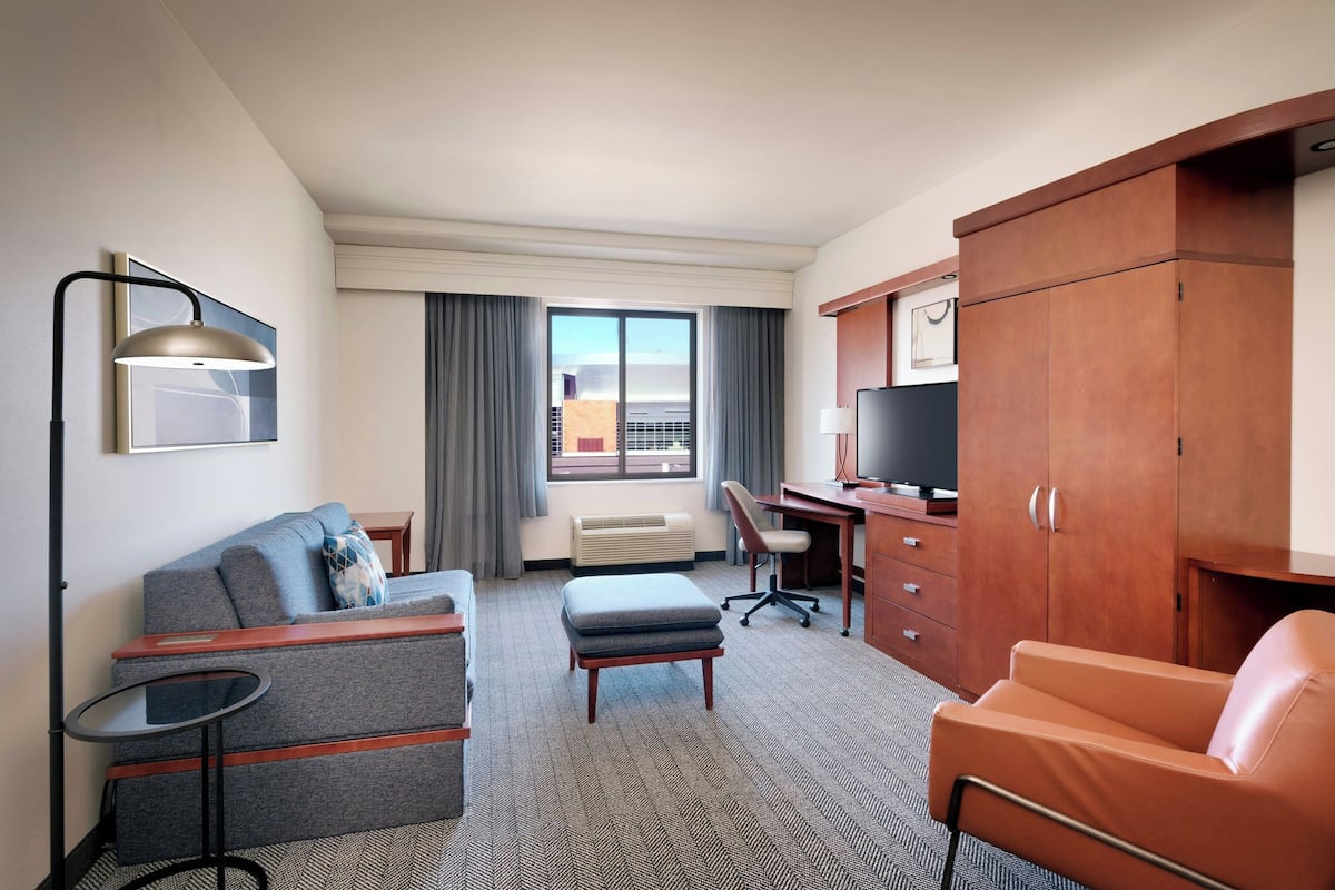A hotel room with a sofa, ottoman, desk, chair, TV, wardrobe, and a window with curtains.