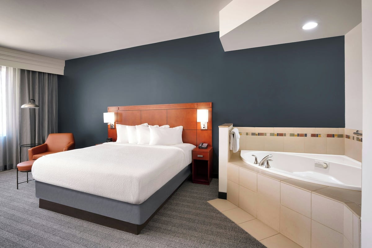 A hotel room with a large bed, a wooden headboard, a bedside table with a lamp, a chair, a bathroom with a bathtub, and a window with curtains.
