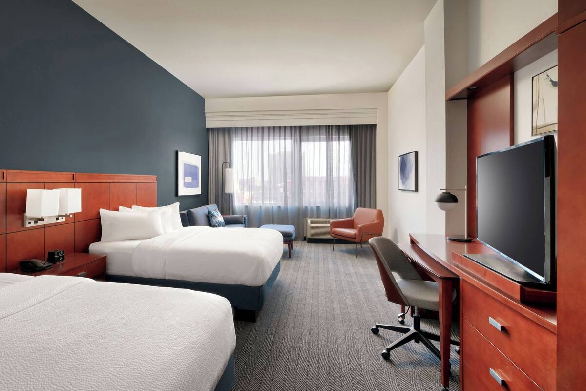 A hotel room with two beds, a desk, a television, and a window with curtains.