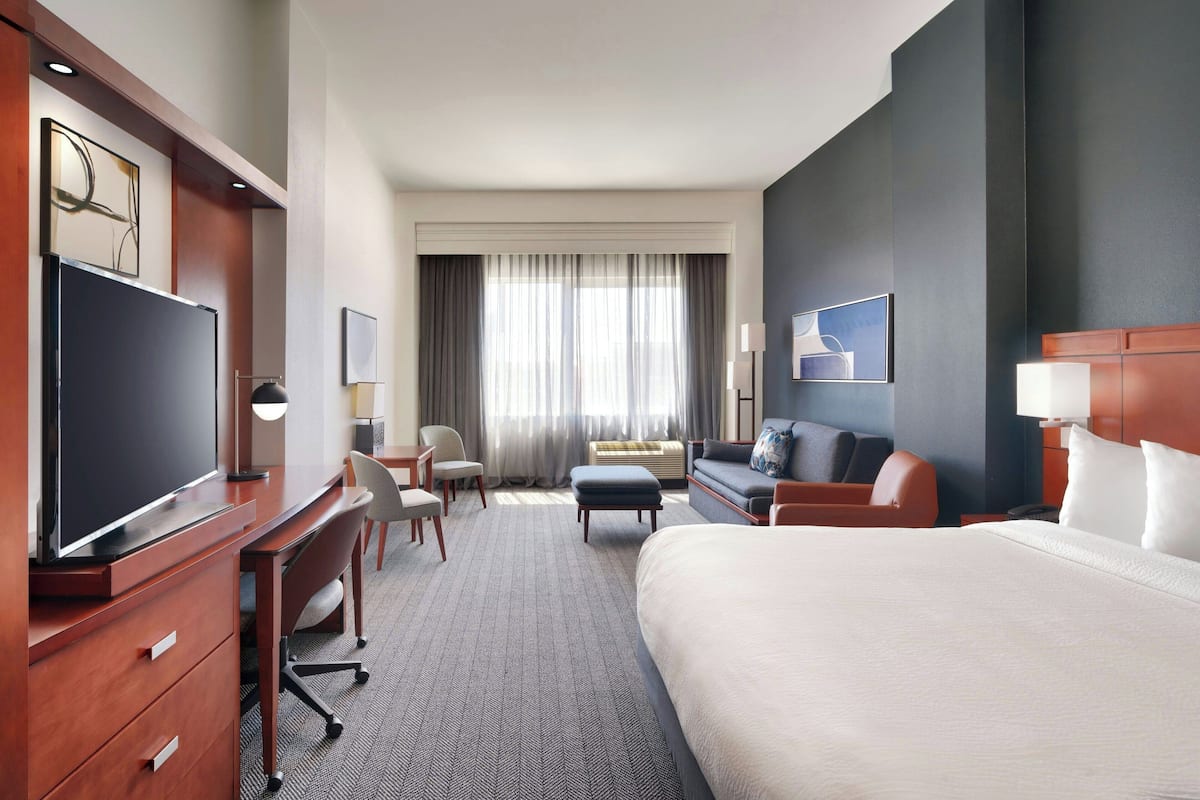 A hotel room with a large bed, a desk with a chair, a sofa, a television, and a window with curtains.
