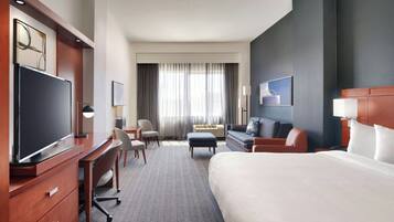 A hotel room with a large bed, a desk with a chair, a sofa, a television, and a window with curtains.