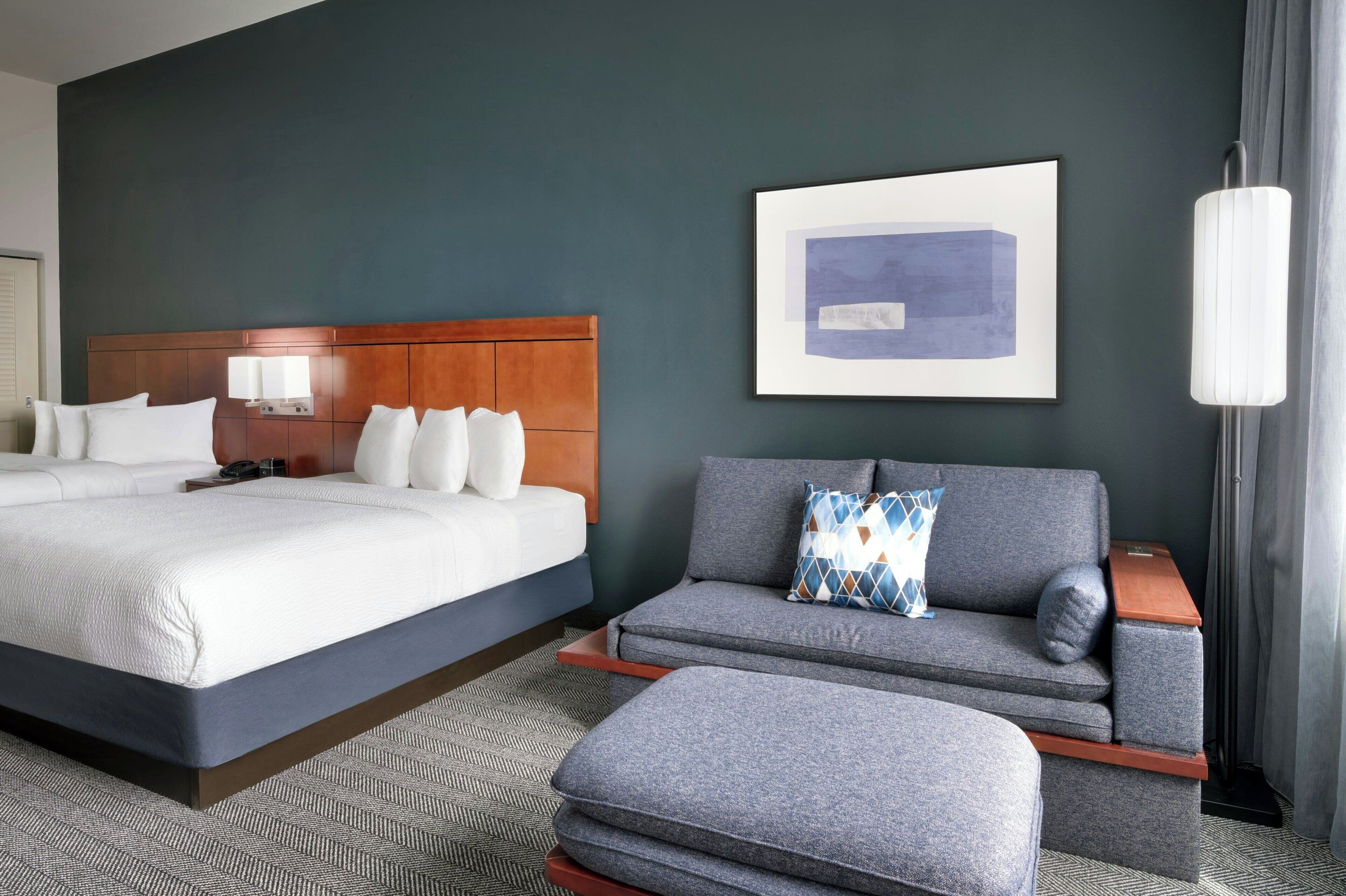 Ein Hotelzimmer mit einem großen Bett, einer Couch und einem Hocker. An der Wand hängt ein gerahmtes Bild und ein Nachtlicht steht daneben.