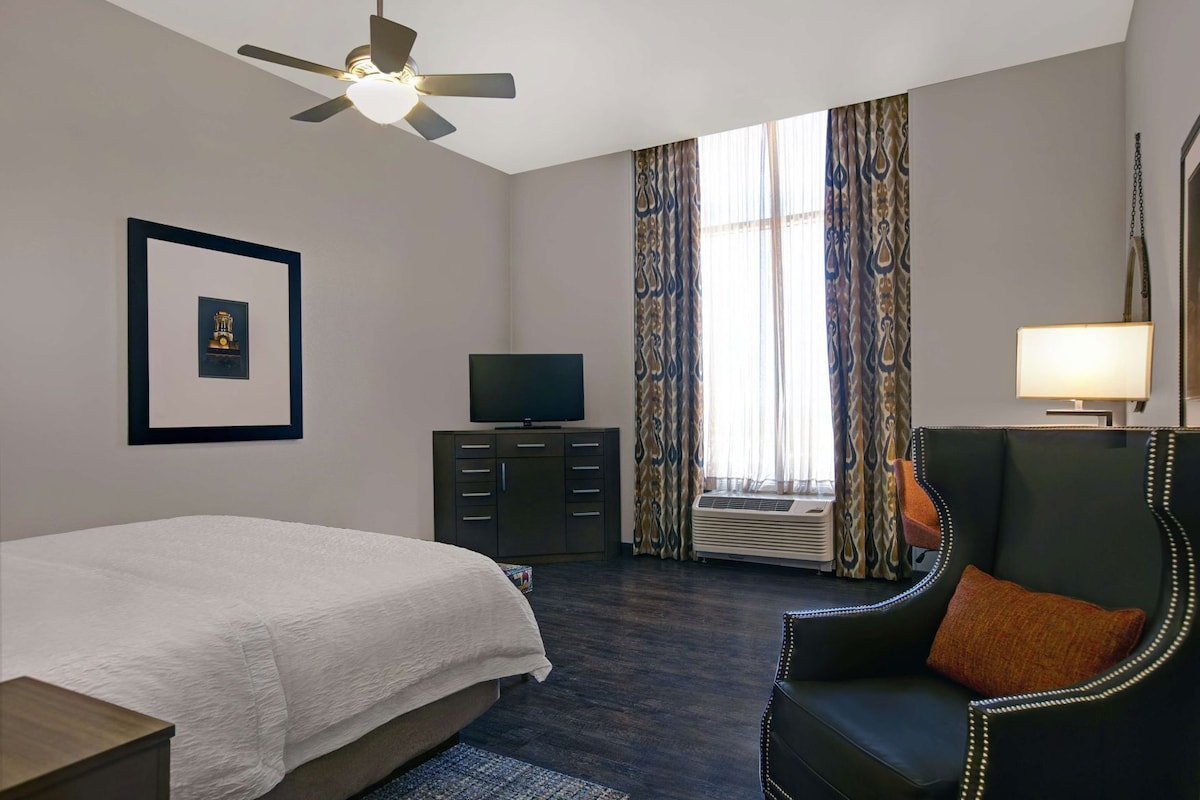 A hotel room with a bed, a television, a desk, a chair, a ceiling fan, and a window with curtains.