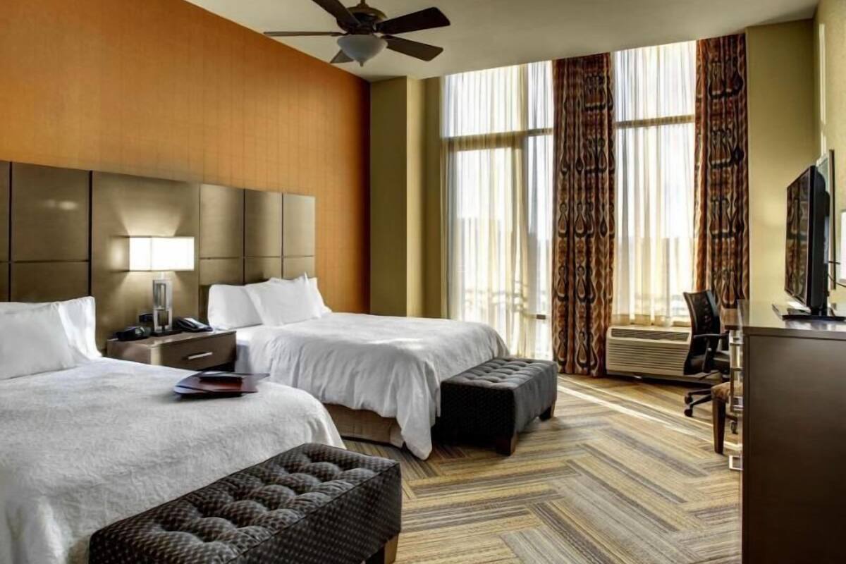 A hotel room with two beds, a desk, a chair, a television, and a ceiling fan.