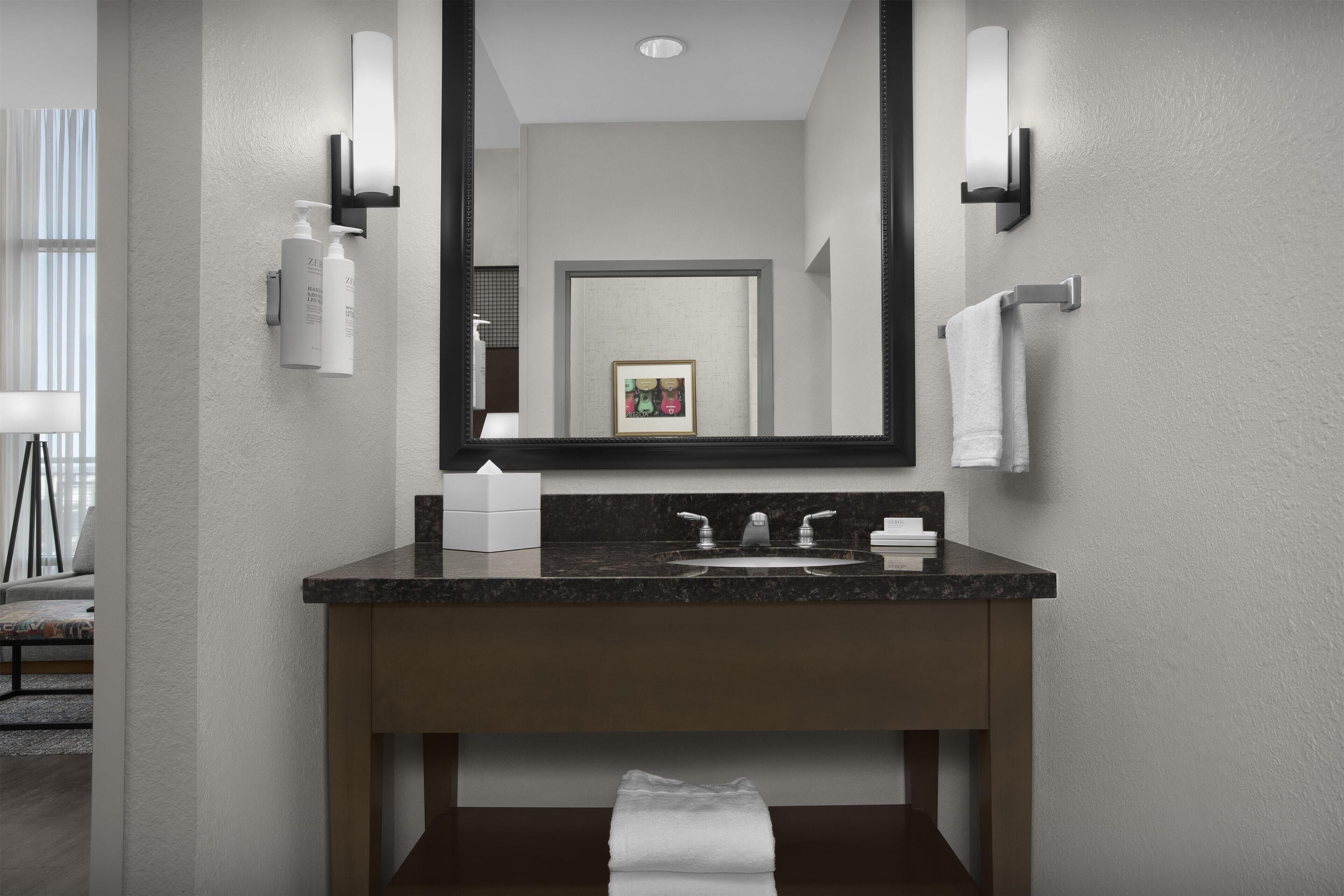 A modern bathroom with a large mirror, a dark granite countertop, a sink with two faucets, and a towel rack.
