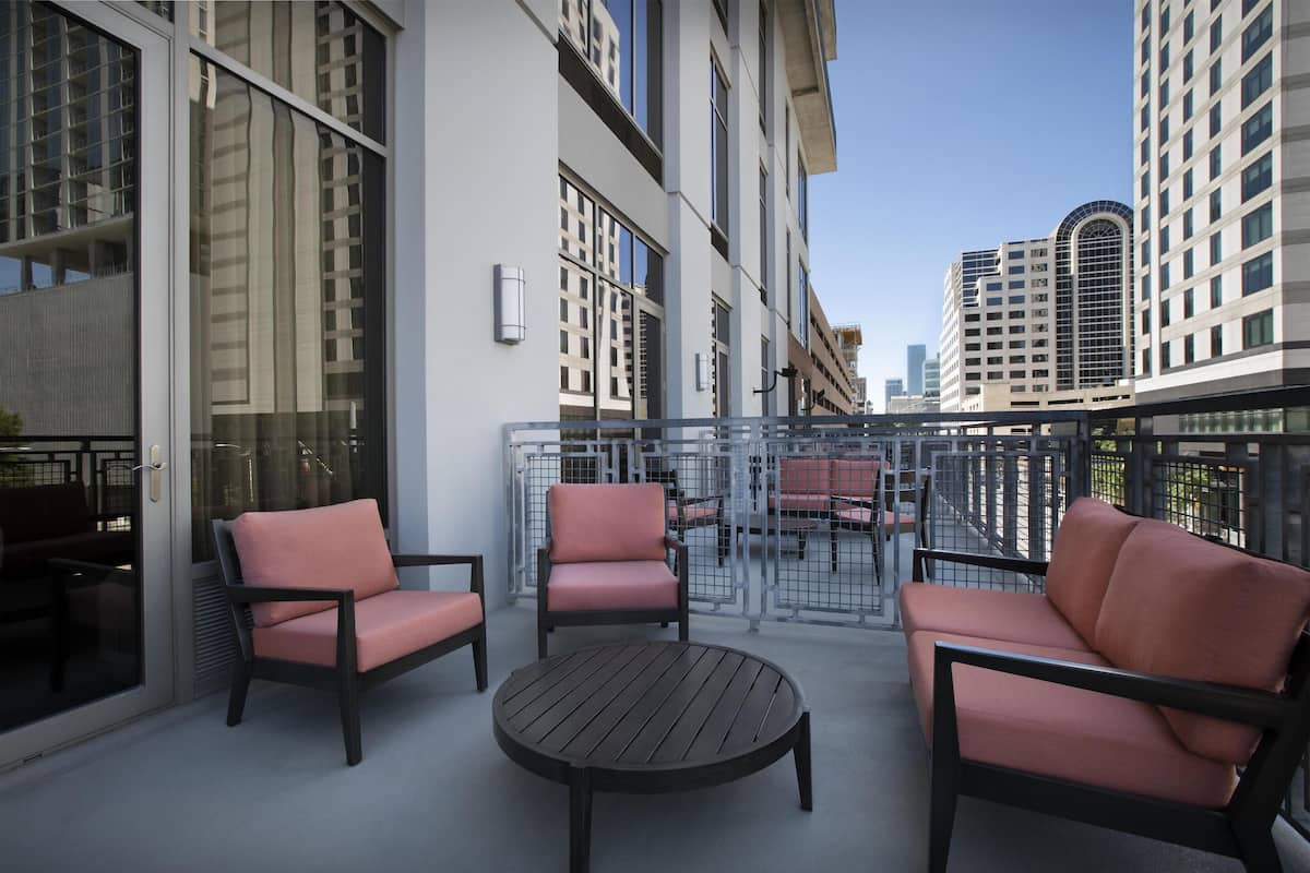 A balcony with two pink cushioned chairs and a round wooden table, overlooking a cityscape with modern buildings.