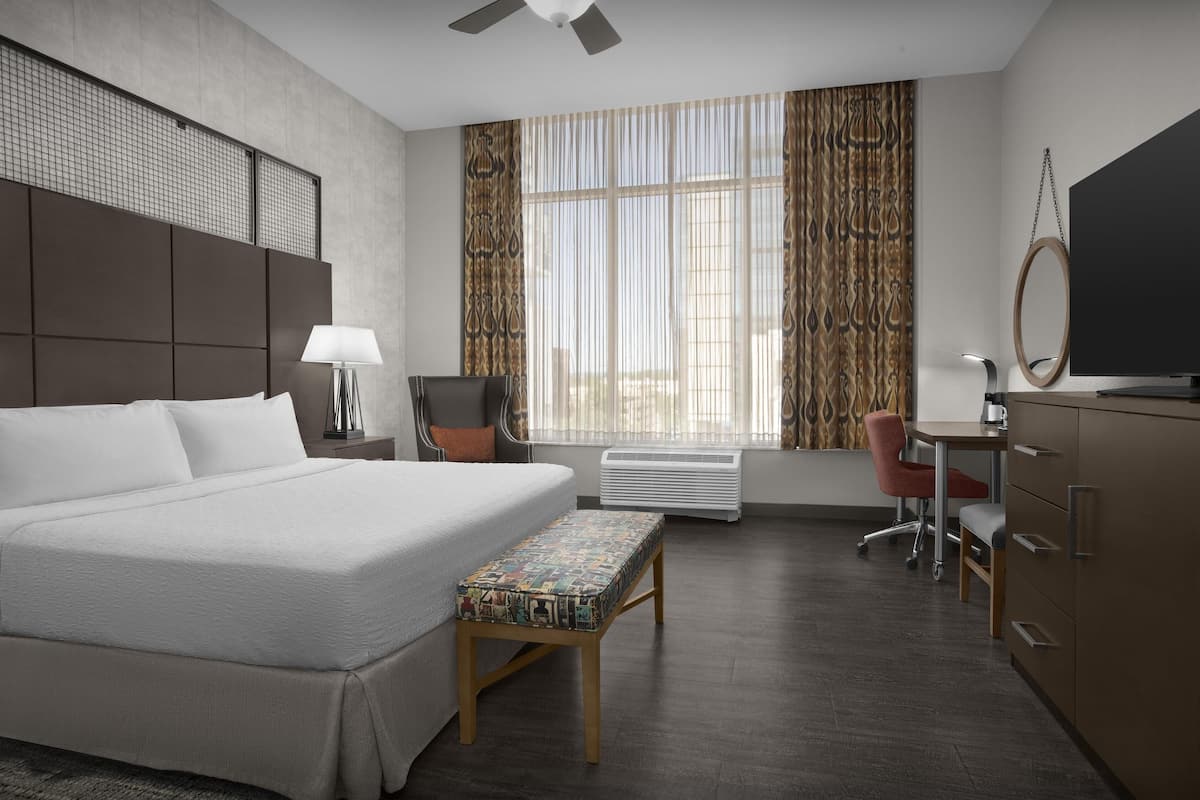 A modern hotel room with a large bed, a desk, a chair, a television, and a window with curtains.