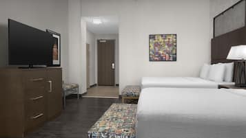 A hotel room with a flat-screen TV, a bed with white bedding, a bench with colorful cushions, a ceiling fan, and a wall-mounted artwork.
