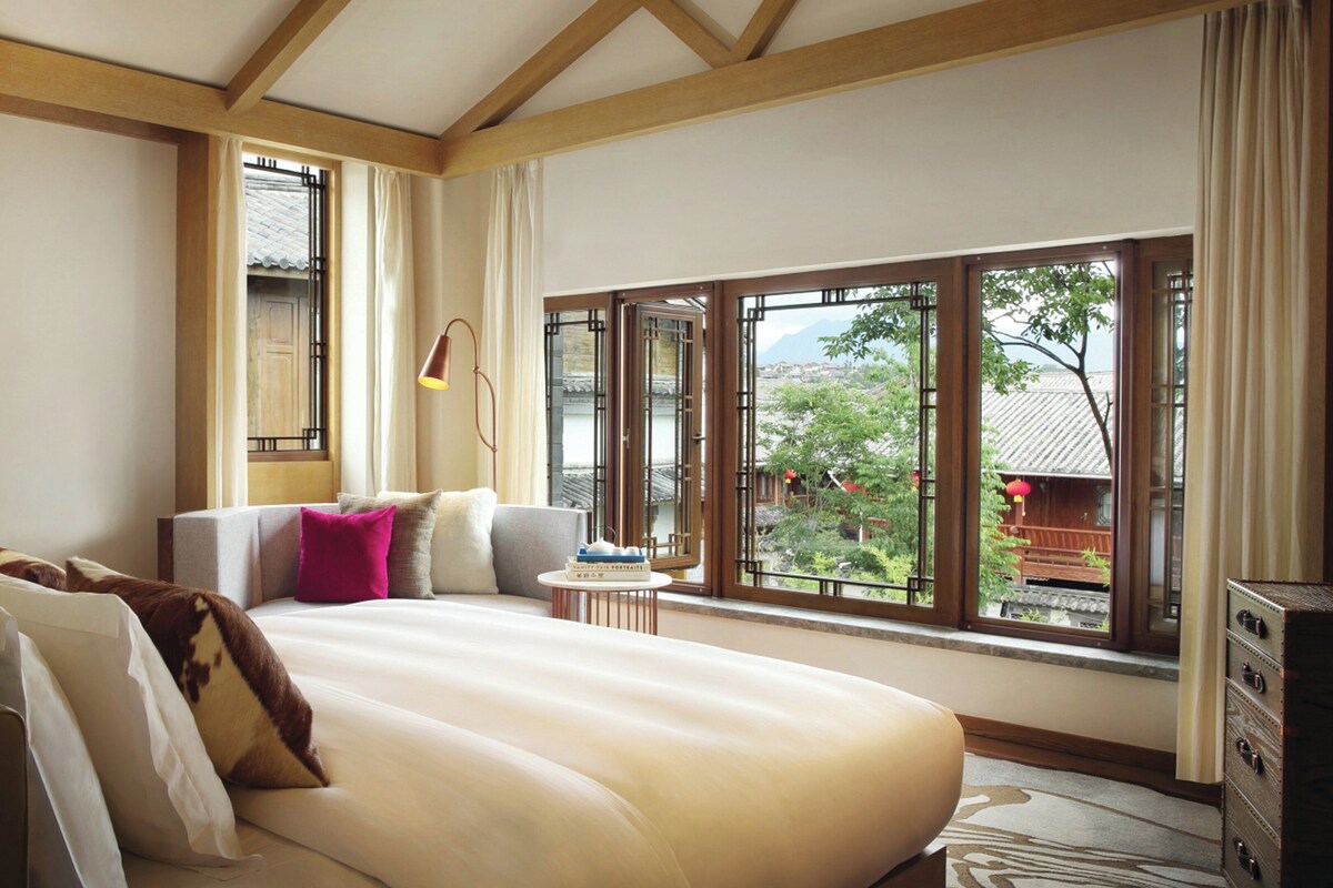 A bedroom with a large bed, a bedside table, a lamp, and a view of the outside.