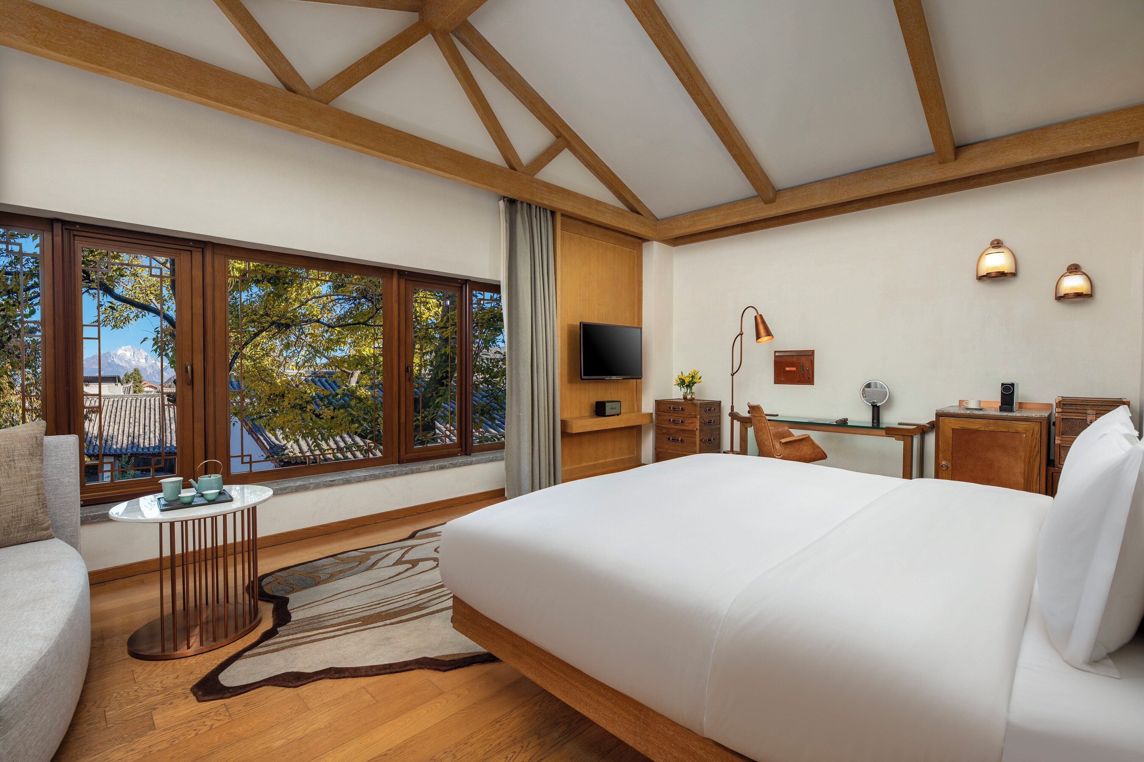 A modern hotel room with a large bed, wooden beams, a TV, a desk with a chair, and a view of trees through large windows.