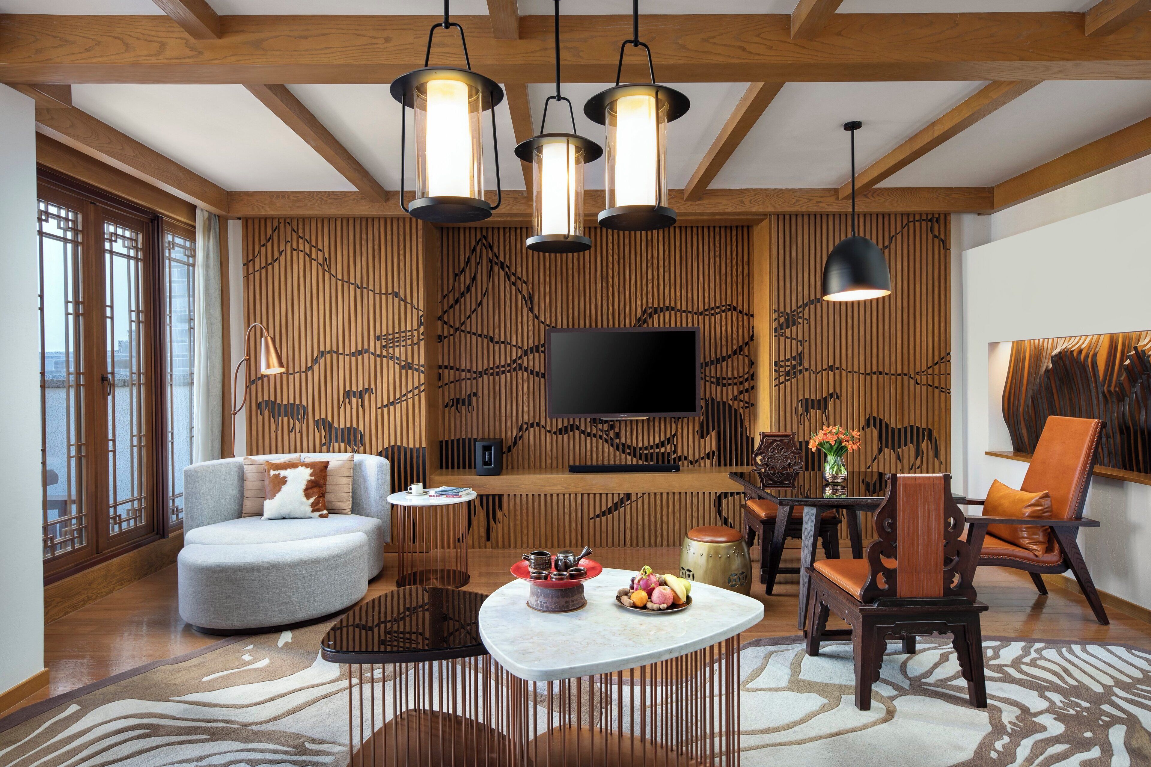 A modern living room with a curved sofa, a round coffee table, and a TV mounted on a wooden wall. There are hanging pendant lights, a wooden floor, and a patterned rug.